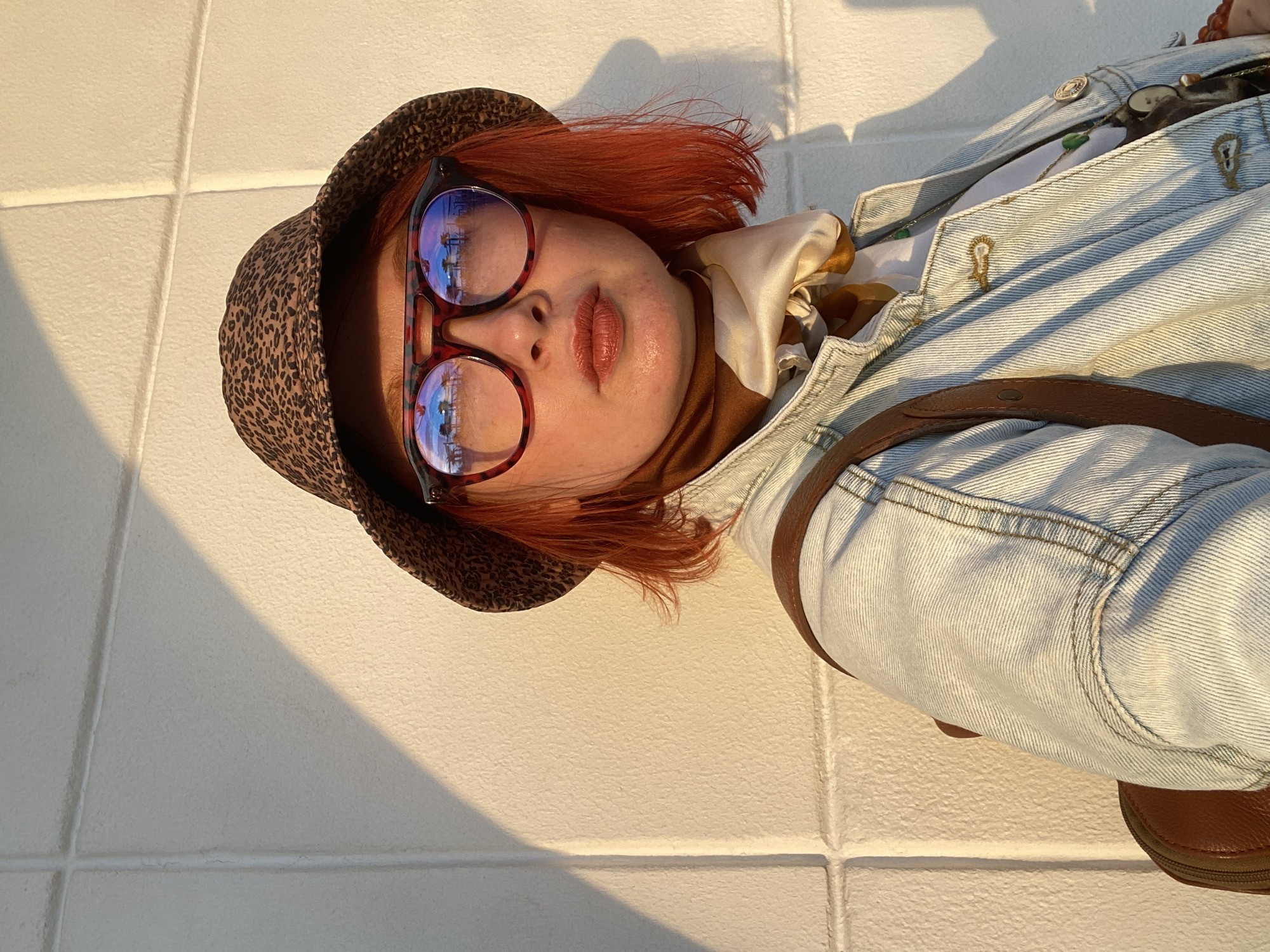 A person with red hair, wearing a leopard print bucket hat and round eyeglasses with red frames, is positioned against a white tiled wall illuminated by strong sunlight. The eyeglasses reflect a cityscape, possibly a coastal view of Sochi, Russia. The person wears a brown turtleneck, a patterned scarf, and a light-wash denim jacket with visible buttons and a brown leather strap. A brown leather shoe is visible at the bottom right corner. The tiles on the wall are uniformly arranged and partially shadowed.
