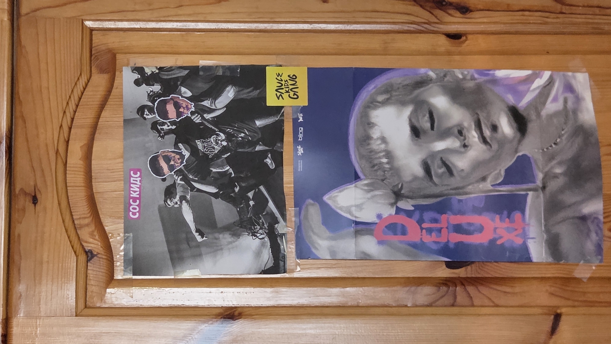 This is a close-up, eye-level, medium shot photograph of two posters affixed to a wooden door. The door appears to be made of varnished pine wood with vertical planks and some visible knots and grain. The top poster is a black and white image of a group of people, possibly musicians, in what looks like a live performance setting. Several individuals are visible, some with sunglasses. A pink rectangular label with Cyrillic text, "СОС КИДС" (SOS KIDS), is adhered to the top of this poster. To the right of the bottom poster, a small yellow sticky note with handwritten text, "SAUCE KIDS GANG", is attached to the door.
The bottom poster features a stylized, grayscale portrait of a person with eyes closed, adorned with a flower or plant arrangement on their head. Pink Cyrillic text, "DELUXE", is vertically aligned on the left side of this poster. Small logos, including "Saice", "bling", and "NTT", are visible in the top right corner of the bottom poster. The posters are taped to the door with clear tape. The scene is indoors, and no specific city or location is discernible from the image itself.