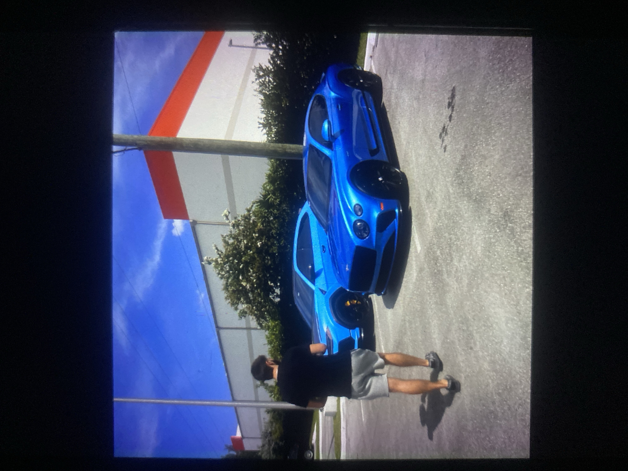 A man is standing in a parking lot, looking at two blue luxury cars. One is a Lamborghini Urus SUV, and the other is a Bentley Continental GT. The man is wearing a black t-shirt and gray shorts. The cars are parked in front of a building with a white and orange facade, under a clear blue sky with some wispy clouds. There are trees and bushes in the background.