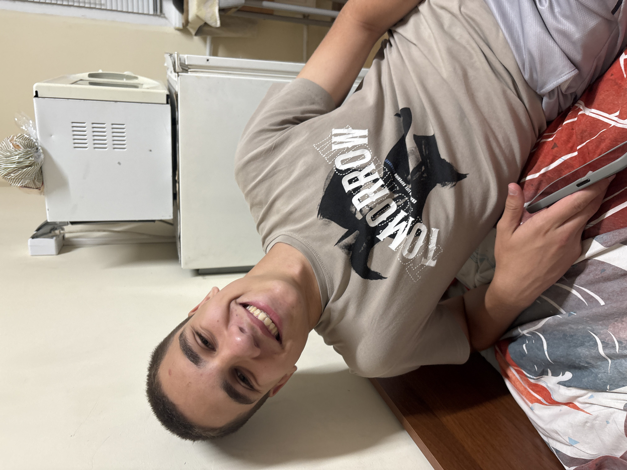A young male, appearing to be in his late teens or early twenties, is depicted reclining on a bed in what appears to be a residential room in Ruse, Bulgaria. He has a short buzz cut and is smiling broadly, revealing both upper and lower teeth. He is wearing a light tan t-shirt with a dark graphic print that includes the white text "TOMORROW" and smaller text "ERA LIFE SUPERIOR STANDARD." He is also wearing light grey shorts. His left arm is bent, resting on the patterned duvet, while his right hand holds a dark grey smartphone. The bed features a wooden headboard and a duvet with a multi-colored pattern in shades of red, blue, grey, and white. Behind him and to his left is a plain, light-colored wall. To his right, a white, two-door refrigerator is visible, topped by a white microwave oven. The microwave has vertical ventilation slits on its left side. A plastic-wrapped, striped item, possibly a loaf of bread, rests on top of the microwave. Further to the right, a partial view of a window with white Venetian blinds is discernible, alongside a section of a light-colored wall and what appears to be a small table or shelf.