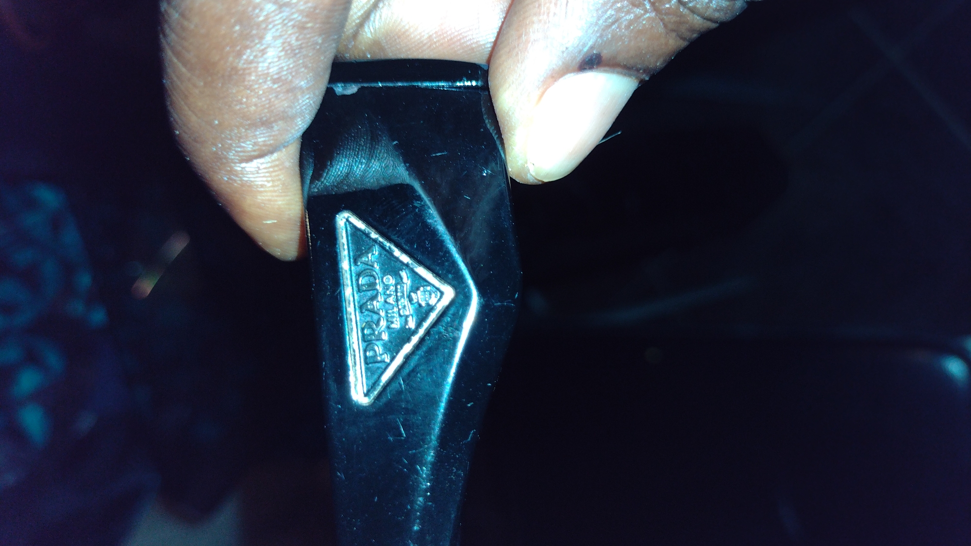 The image shows a close-up of a black object, likely sunglasses, with a metallic Prada logo embedded on it. A person's fingers are holding the object, obscuring part of it. The background is dark and out of focus, with some faint blue and teal lights visible. The overall scene is dimly lit, suggesting it was taken in low-light conditions. There are no discernible details indicating a specific location within the city of Jos, Nigeria.