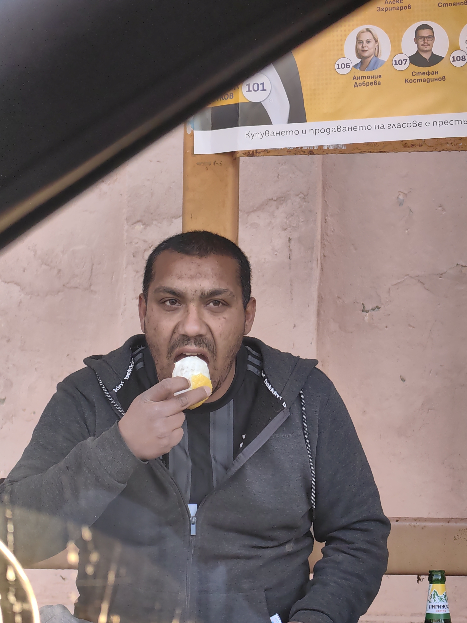 A medium eye-level shot captures a man eating. He is dark-haired with a dark mustache, wearing a dark gray hoodie and a black t-shirt. He is holding a small yellow and white item to his mouth, apparently eating it. Behind him, a light peach-colored wall displays a political advertisement with three headshots of candidates, numbered 106, 107, and 108, alongside the number 101. Text in Bulgarian accompanies the images. A green beer bottle labeled "ПИРИНСКО светло-" is visible on the bottom right. The scene is possibly in Hirevo, Bulgaria, based on the Bulgarian text.