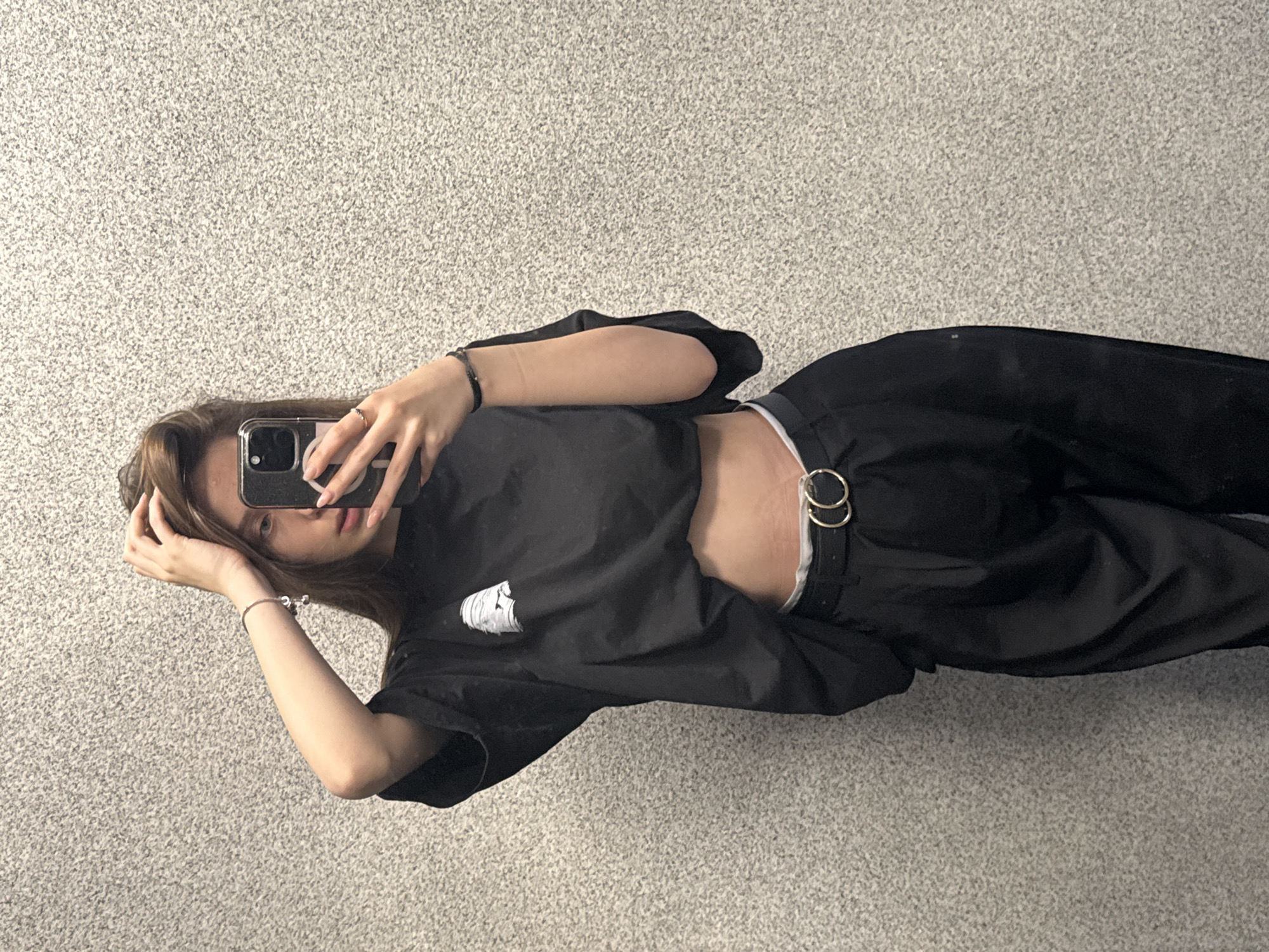 A young woman is posing for a selfie in front of a light gray textured wall. She is wearing a black crop top and black pants with a belt that has two large metal rings. She is holding a smartphone in her right hand to take the photo, while her left hand is touching her hair. She is standing indoors. Without additional information, it is impossible to determine if the location is in Sofia, Bulgaria.