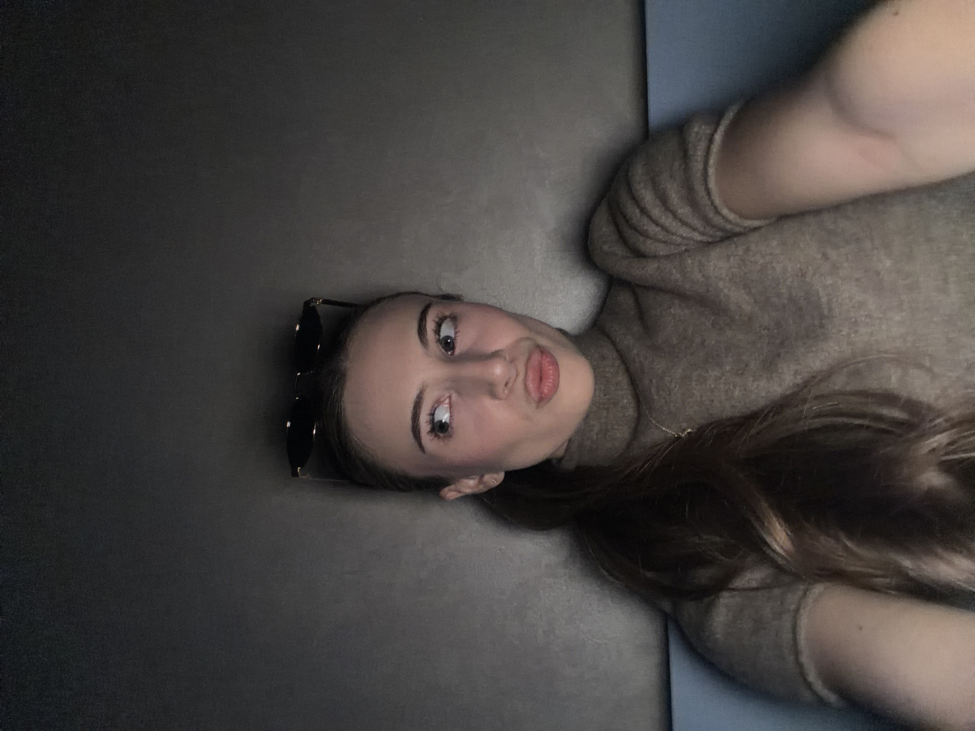The image displays a young adult female captured in a selfie from the chest up, positioned indoors against a plain dark grey wall. A thin light blue surface is visible along the lower frame. She has long, dark brown hair pulled into a ponytail, with dark-framed sunglasses perched on top of her head. She wears a short-sleeved, ribbed turtleneck garment in a muted brown color and a fine necklace chain. Her gaze is directed upwards and to her left, with her lips puckered. The lighting is subdued. This scene is situated in Plovdiv, Bulgaria.