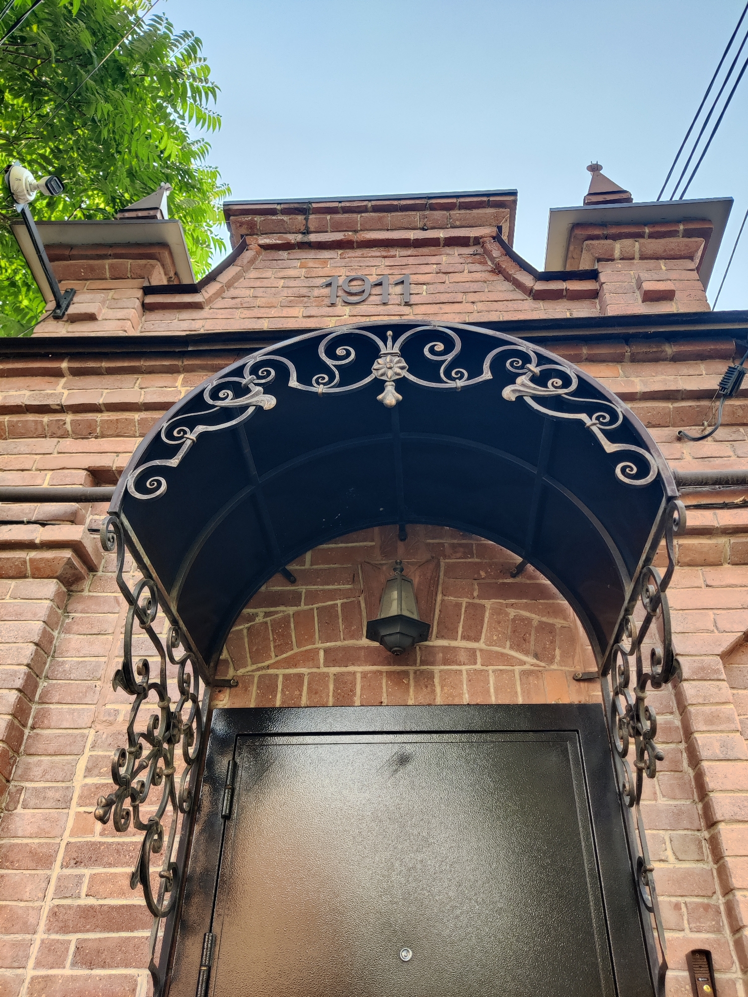 This is a close-up, low-angle shot of a brick building entrance. The primary subjects are a black metal awning with ornate scrollwork, a black metal door with a textured finish, and a vintage-style lamp hanging from the awning. The building facade is constructed from red brick, with decorative brickwork at the top of the entrance. Above the awning, the number "1911" is visible in dark metal lettering, suggesting the building's construction year.
The scene is set outdoors, with a bright, clear sky visible at the top of the frame, indicating daytime with fair weather. To the left, a lush green tree branches into the frame, adding a natural element to the urban architecture. A security camera is mounted on a pole to the left of the building, and electrical wires are strung across the sky at the top right. The overall impression is one of historical architecture with some modern security features. There are no people visible in the frame. The setting appears to be a street or alleyway in a historical part of Krasnodar, Russia, given the architectural style and the date inscribed on the building.