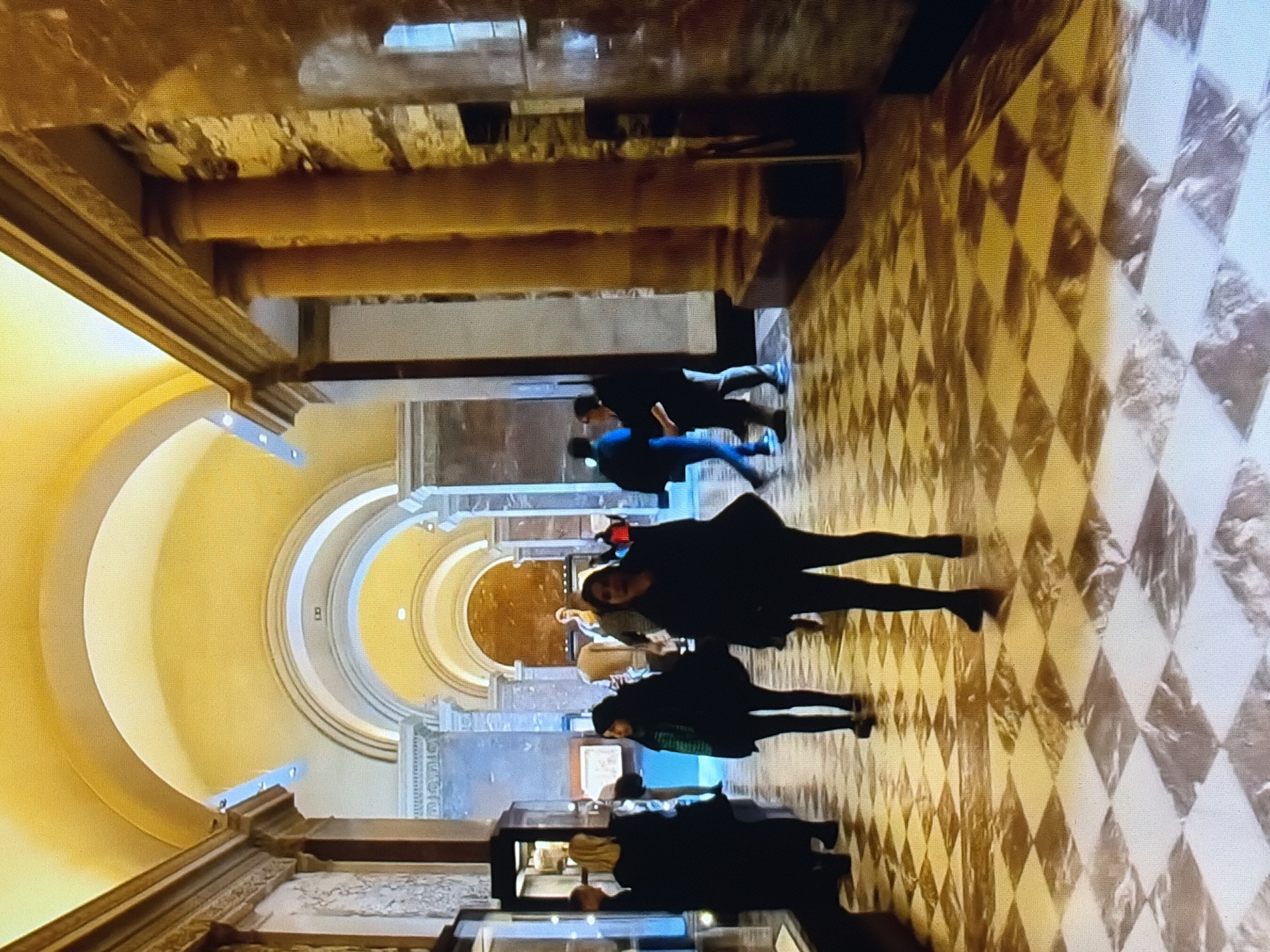 A long, arched corridor in a museum or grand building is depicted. The walls are adorned with classical architectural elements, including columns and intricate carvings. The ceiling is a smooth, golden arch. The floor is a checkered pattern of marble tiles, with a glossy finish that reflects the ambient light. Several people are present, walking through the corridor. Some are looking at displays along the walls, while others are engaged in conversation. The lighting is warm and somewhat dim, casting long shadows. The overall atmosphere suggests a place of historical significance and cultural interest.