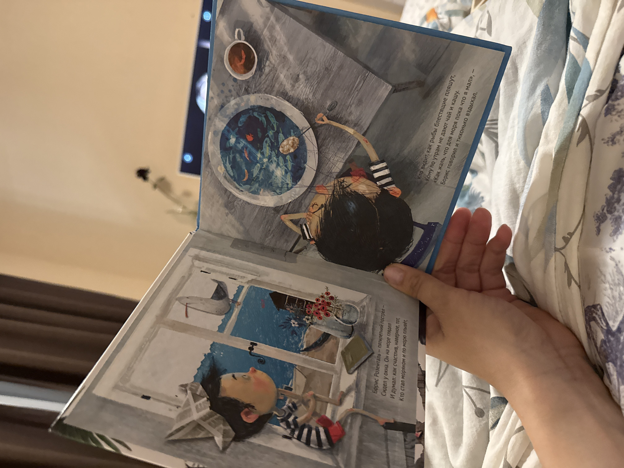 The image shows a person holding open a children's book with colorful illustrations. The book appears to be written in Russian, with text visible on both pages. One illustration depicts a child sitting at a table, looking into a bowl filled with fish, while a cup of tea sits nearby. The other illustration shows a child lying by a window, gazing out at a seascape. The book is being held against a background of patterned bedding. There is no indication of location in Tobolsk, Russia in the photo.