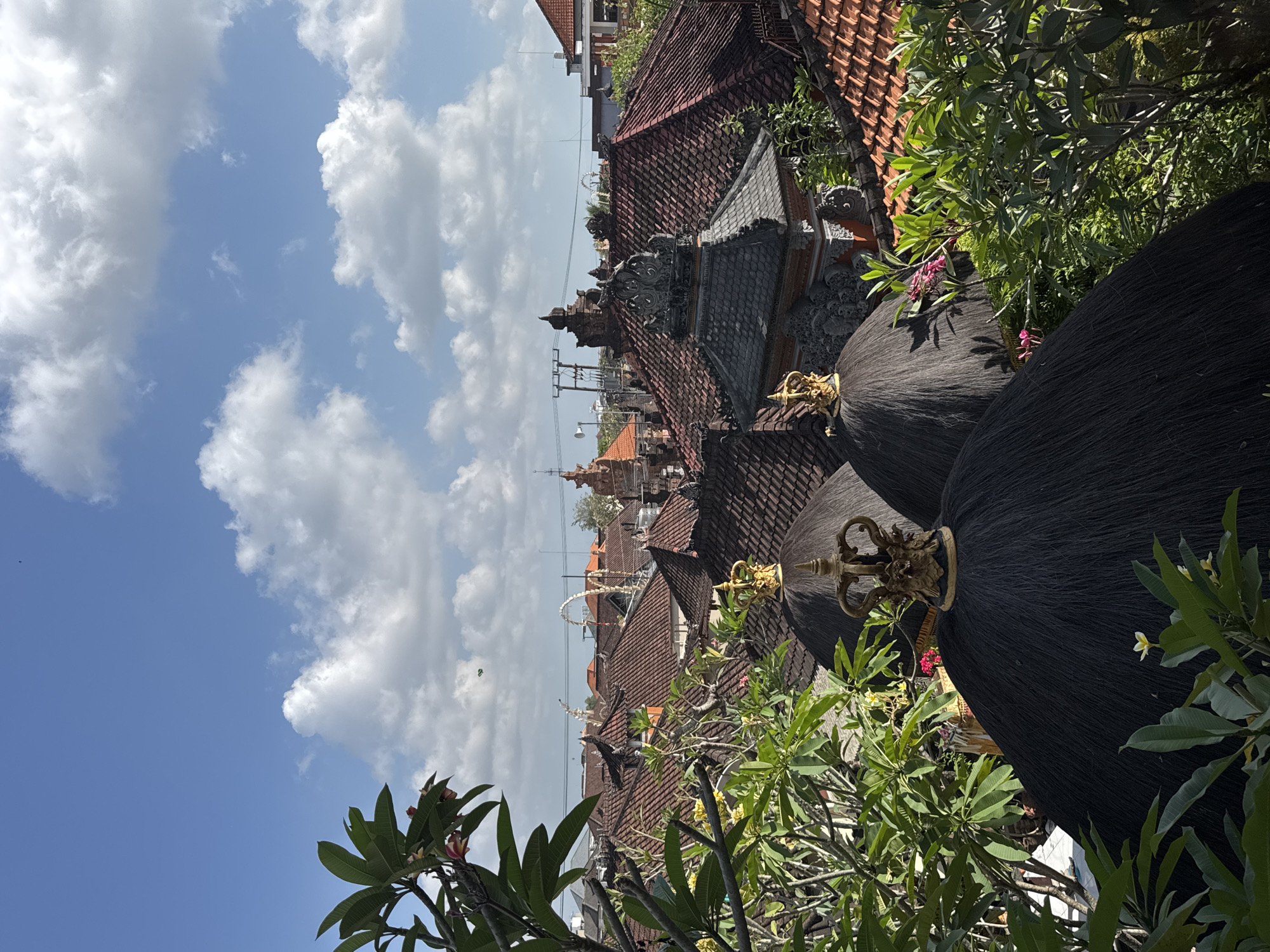 A high-angle view shows a cluster of traditional Balinese buildings with dark, thatched roofs and terracotta tile roofs under a blue sky with scattered white clouds. The foreground is dominated by two large, rounded, dark thatched structures topped with ornate gold finials. Lush green foliage with visible frangipani trees and pink flowers partially obscures the structures and extends into the mid-ground. Behind the thatched roofs, a dense arrangement of red-tiled roofs covers the buildings. In the distance, more buildings with red tiles and intricate architectural details are visible, along with utility poles and wires. The scene captures the characteristic architecture of North Kuta, Indonesia, bathed in bright sunlight. There are no people visible in the image.