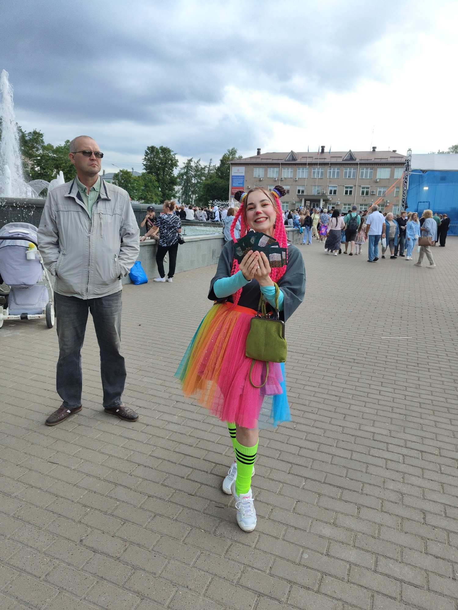 In an outdoor urban setting, a woman dressed as a clown with bright pink braided hair and a multi-colored tutu stands with a smile, holding several items that appear to be tickets or brochures. She is wearing bright green socks and white sneakers. To her left, a man in a grey jacket and dark jeans stands with his hands in his pockets, wearing sunglasses. Behind them, a fountain spouts water, and a stroller is visible. In the background, a building with multiple windows and flags stands under a cloudy sky, with a crowd of people gathered. The ground is paved with brick-like stones. The scene appears to be a public gathering or event in an urban plaza.