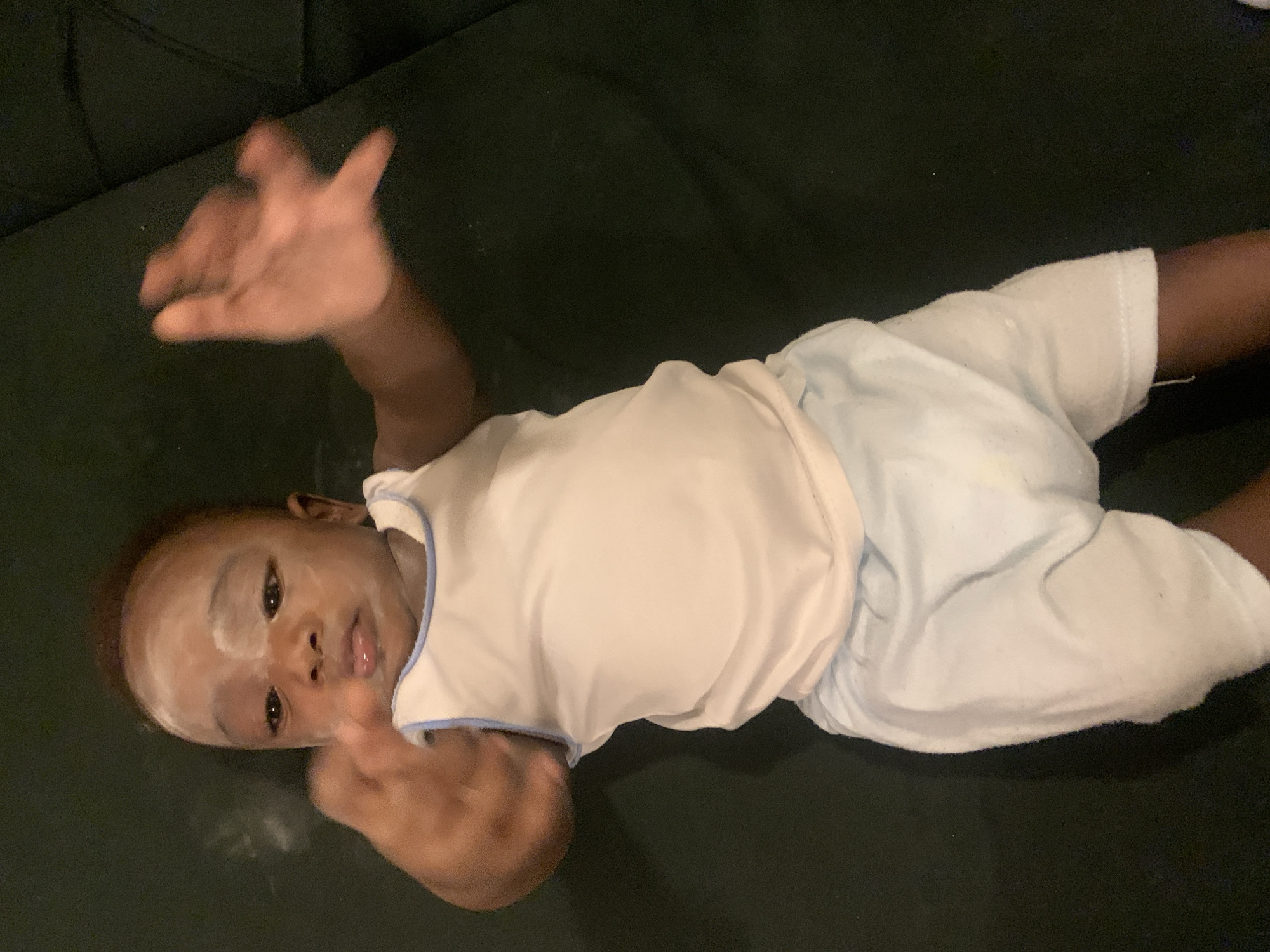 A baby with a white substance on their face lies on a dark surface, possibly a couch. The baby is wearing a white shirt with light blue trim and light blue shorts. One arm is raised with the hand open, while the other is by their side. The location and city information provided is not present in the image.