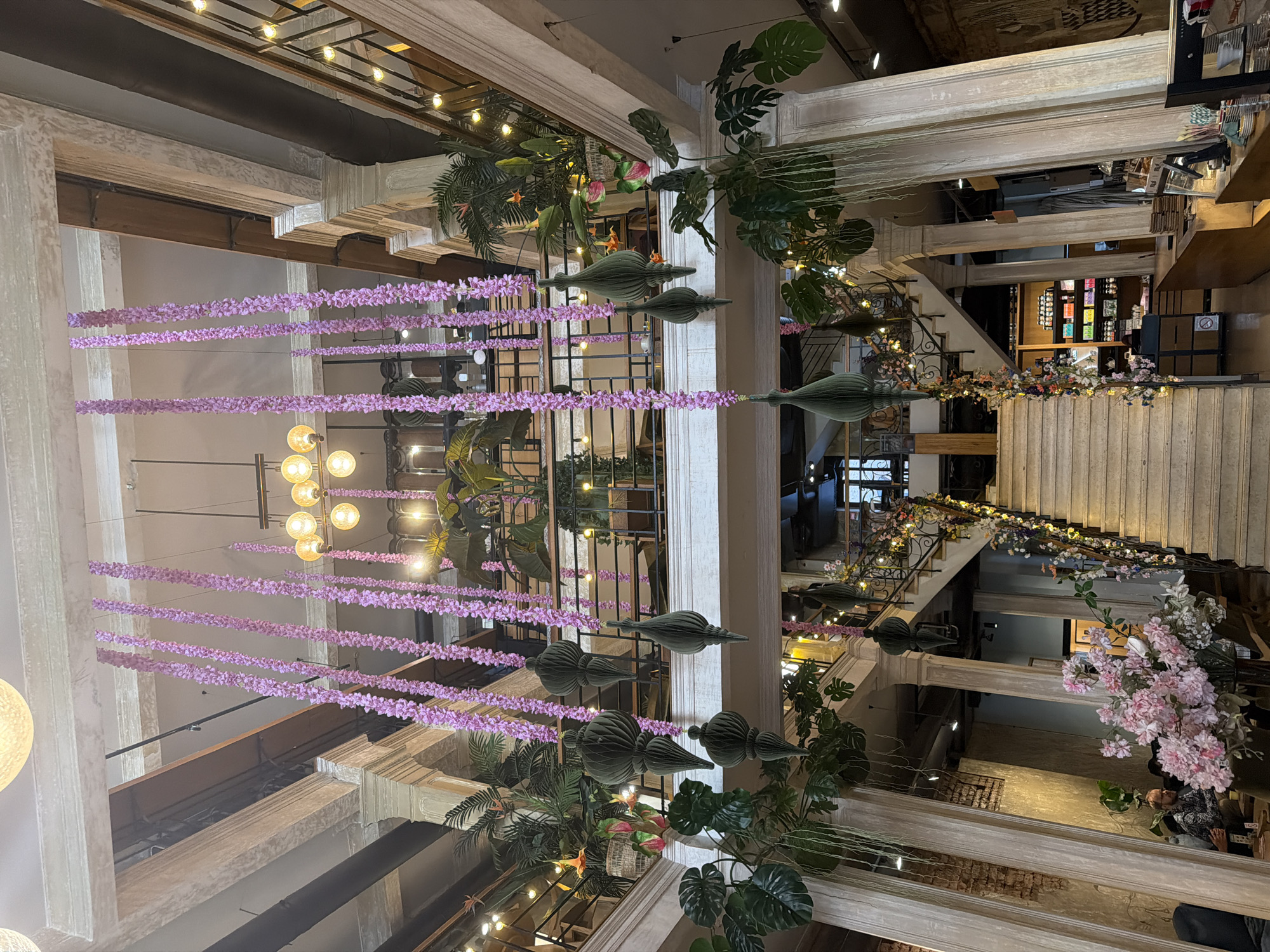 The image shows an interior space in Belgrade, Serbia. It features a multi-level area with columns and staircases. Floral decorations, including strings of purple flowers and arrangements of greenery, hang from the ceiling and adorn the staircases. The space seems to be a retail or commercial area, with merchandise visible on shelves in the background. People are present in the background. The lighting is a combination of natural light and artificial fixtures, creating a warm and inviting ambiance.