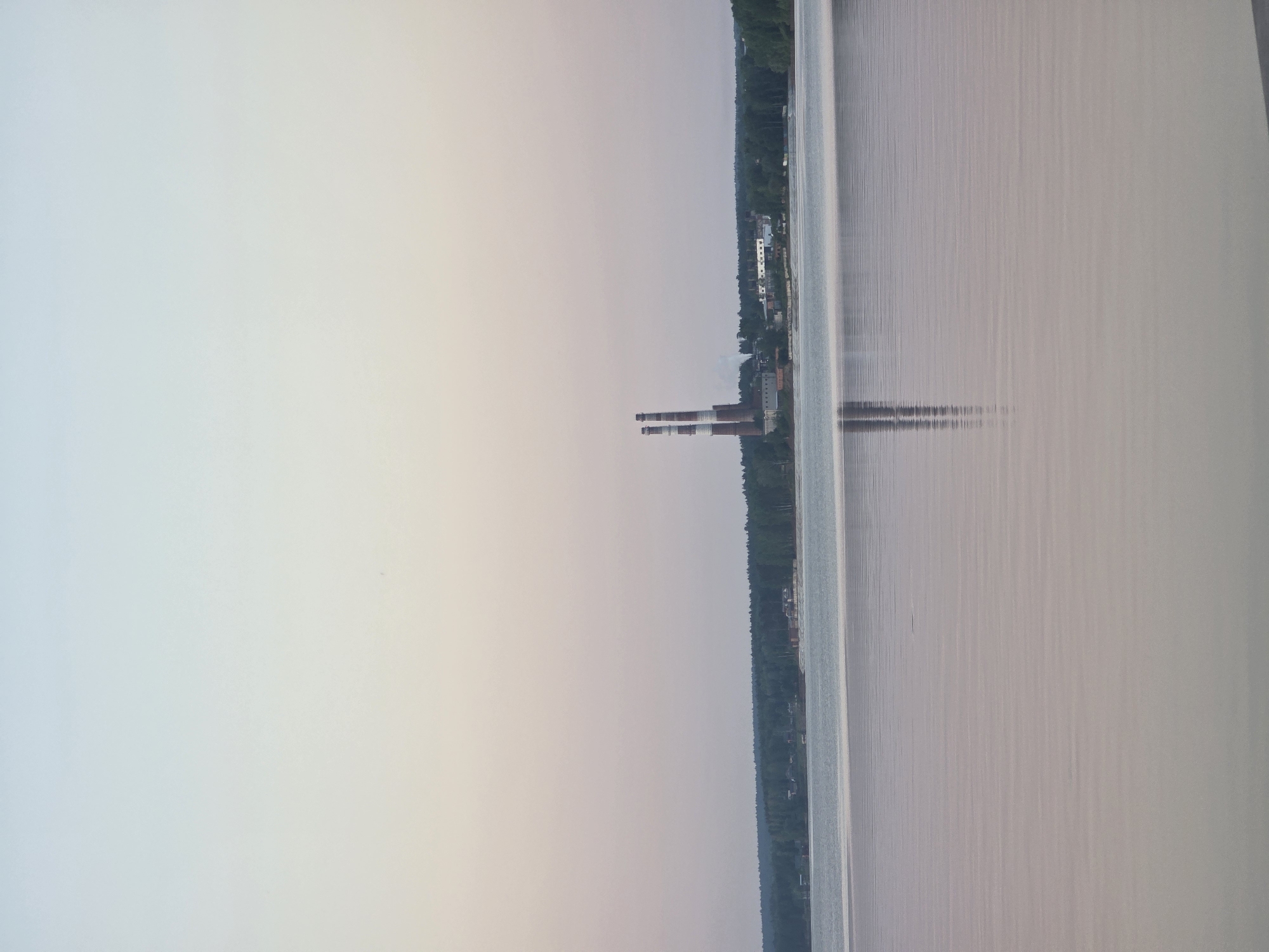 The image depicts a scene in Chaikovsky, Russia, possibly a reservoir or large body of water with a shoreline visible on the right side. The water surface appears calm with subtle ripples. Along the shoreline, there is a dense forest and some buildings with chimneys. The sky is pale and hazy.