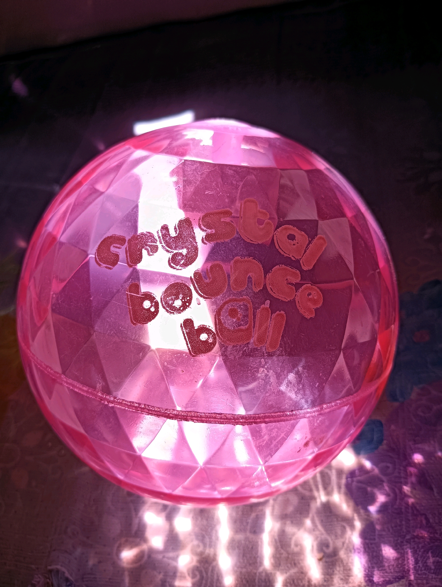 A pink, faceted, translucent ball rests on a patterned surface. The ball has the words "Crystal Bounce Ball" written on it in a darker pink color. The surface beneath the ball appears to be a fabric with a floral or abstract pattern. The lighting is bright, casting a pink glow and reflections onto the ball and the surface. The background is dark and out of focus. There are no people or animals visible in the image. The scene is indoors, with no indication of a specific city or location.