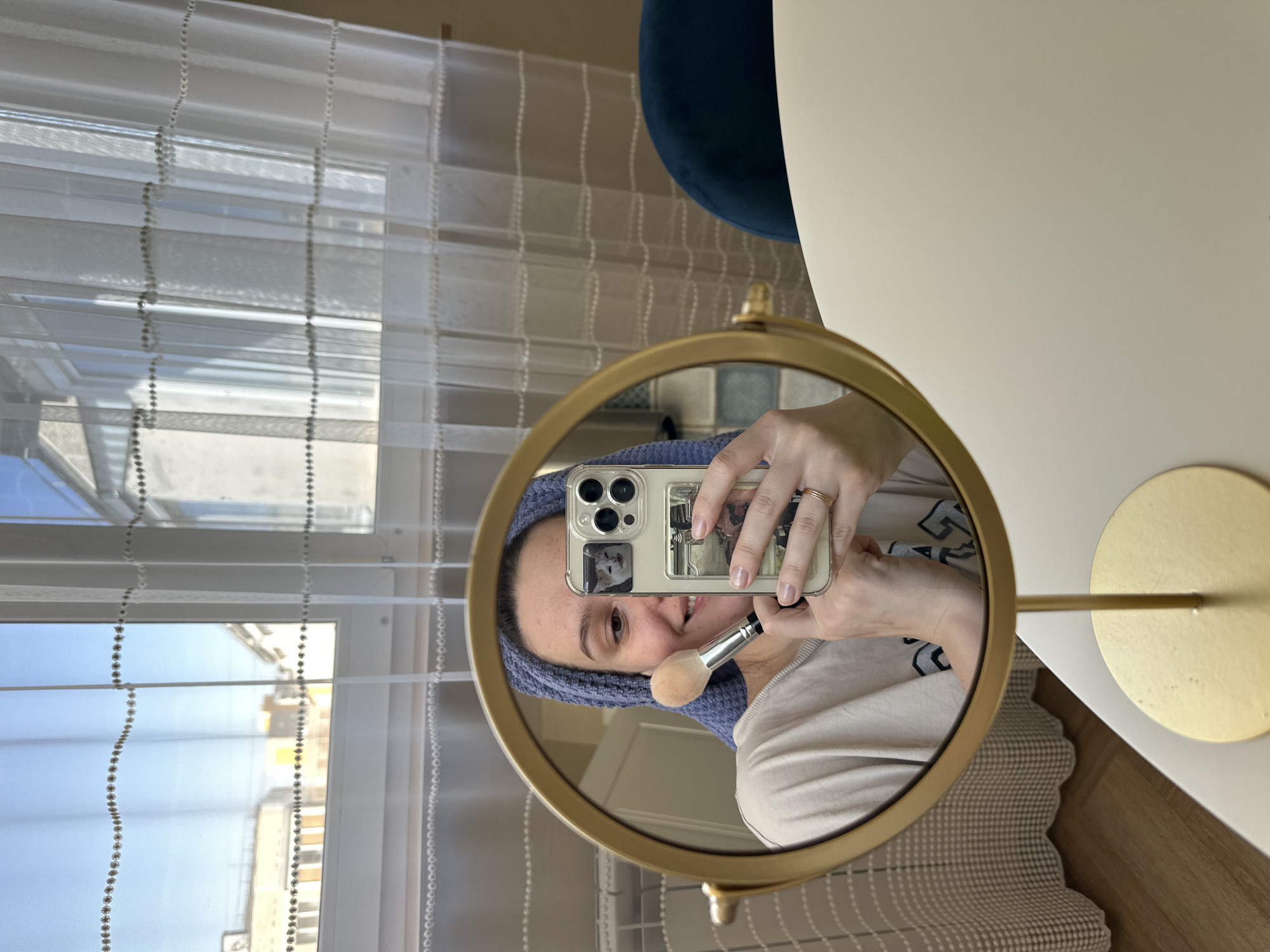 A woman takes a selfie in a round gold-framed mirror. The mirror is on a white table with a gold stand and base. She is wearing a blue towel on her head and holding a makeup brush in one hand and a cell phone in the other. The cell phone is a clear case with an image in the back. The woman is smiling. The background includes a window with white sheer curtains and a view of buildings. A blue chair is partly visible. The location is Chaikovsky, Russia.