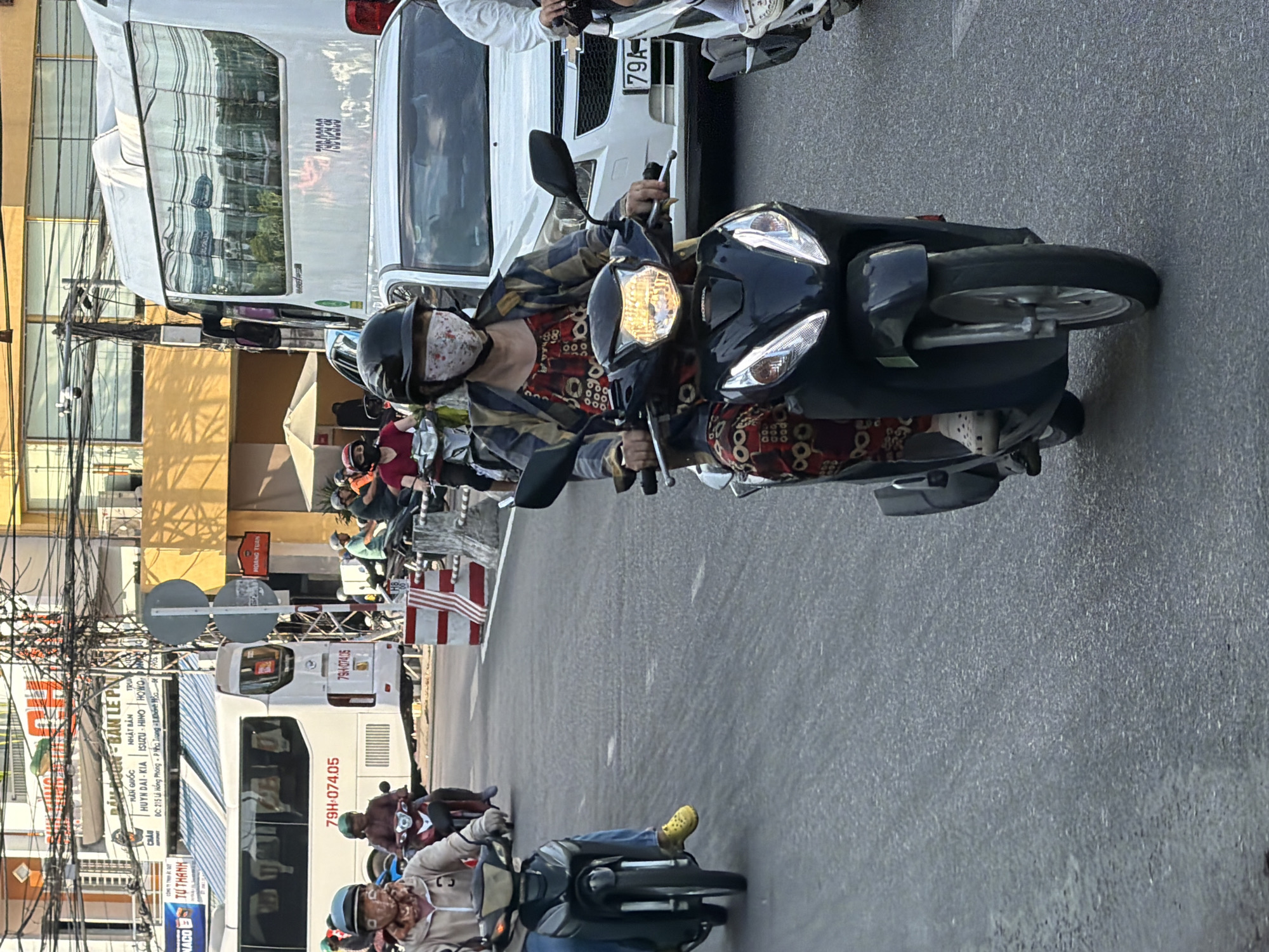 This is a medium shot of a busy street scene in Nha Trang, Vietnam. The image focuses on several individuals riding motorcycles, with a white bus and a white car visible in the background. The foreground features a woman on a black Honda scooter, wearing a floral-patterned mask, a checkered jacket over a red and black patterned dress, and white Crocs. To her left, another person on a motorcycle, wearing a beige jacket and yellow Crocs, is also visible. In the background, a white bus with the license plate "79H-074.05" is seen, as is a white car with a license plate that appears to read "79A...". Traffic cones and signs are present, indicating road construction or a traffic control point. The environment suggests a typical urban setting with overhead power lines and buildings lining the street. The overall impression is one of everyday traffic and transportation in a Vietnamese city.