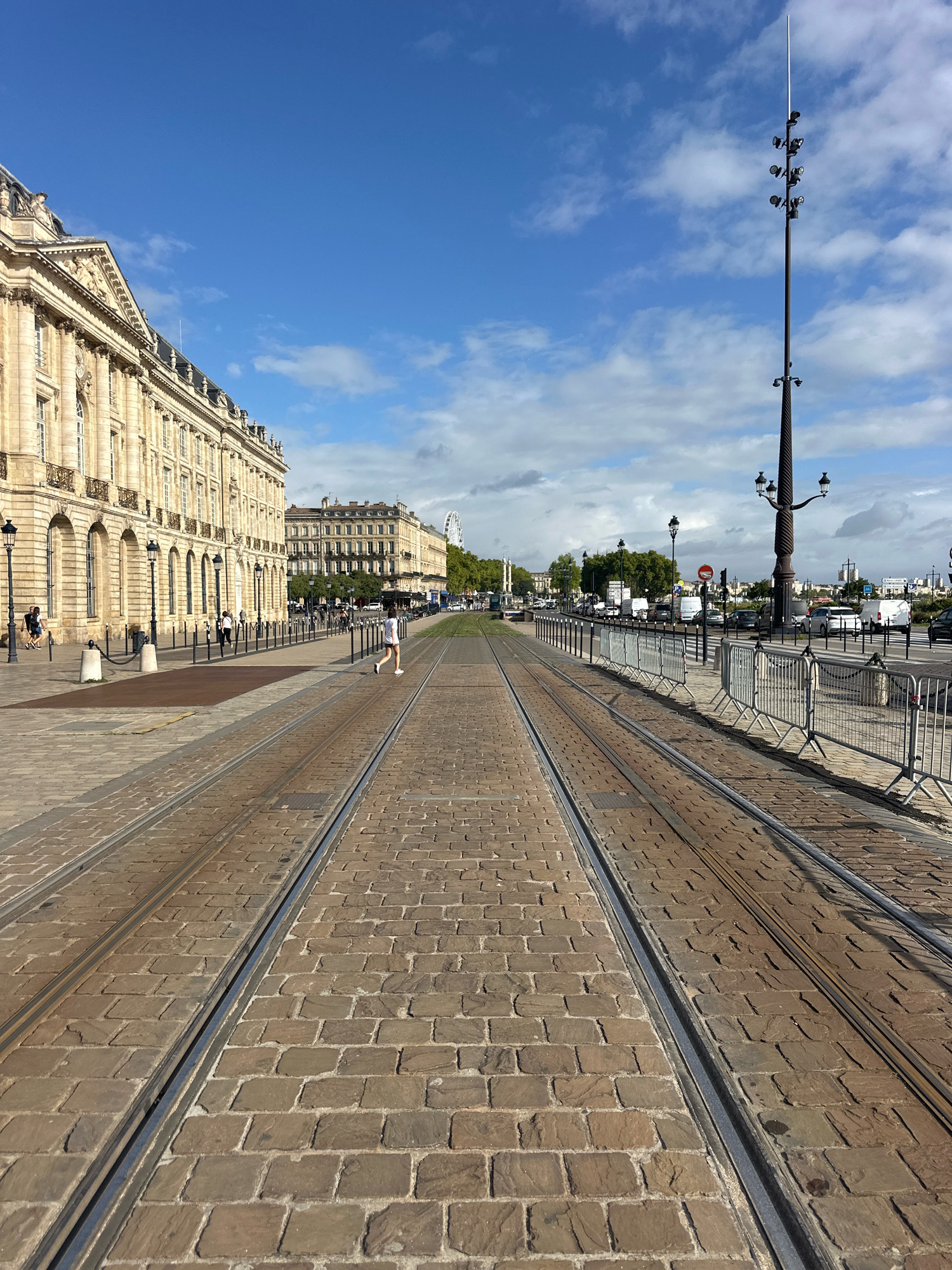This is a low-angle, eye-level shot of a cobblestone street with tram tracks in Bordeaux, France. The street is wide and extends into the distance, with tram tracks forming the central visual element. The buildings lining the street on the left are grand, ornate structures with multiple windows and classical architectural details. In the background, there are more buildings, trees, and a Ferris wheel is visible. The sky is blue with scattered white clouds. Several people are walking or running on the sidewalk and street. There are also cars and vans on the road in the background, and metal barriers are set up along the side of the street. The lighting suggests it is daytime.