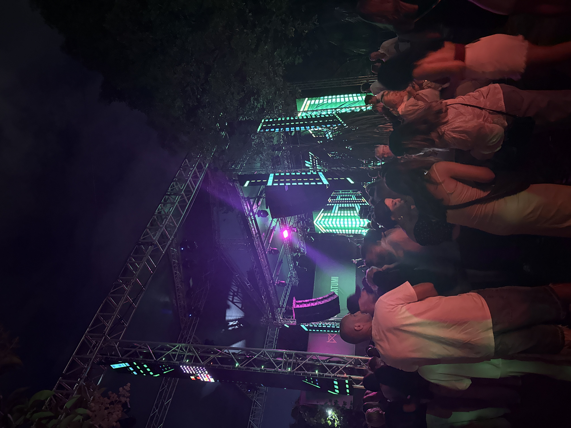 The image depicts an outdoor night-time event in Batumi, Georgia, likely a concert or festival, given the presence of a large stage structure with lighting and sound equipment. A crowd of people is visible in the foreground, gathered to watch the performance. The stage features prominent LED screens displaying a cityscape design with vertical green and blue lights. Trees frame the scene on the right, indicating an open-air venue, possibly a park or public space within the city. The atmosphere is festive, with people dressed casually and appearing engaged with the event. The lighting from the stage and screens casts a colorful glow on the surroundings and the attendees.