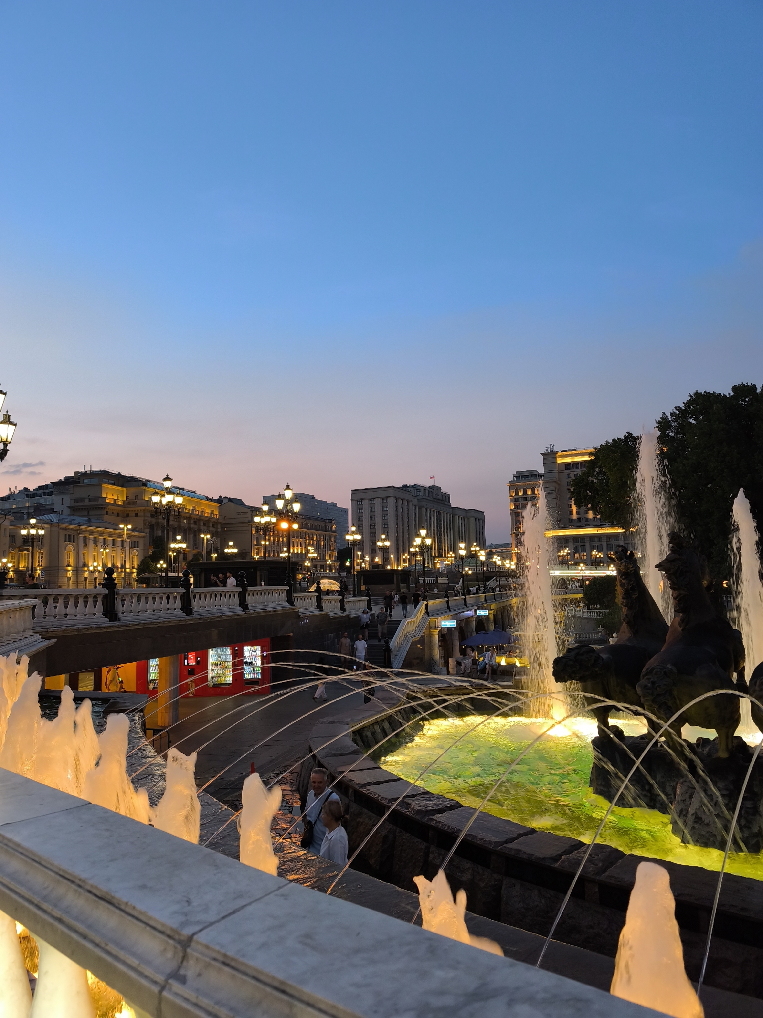 The image depicts a view of a fountain with illuminated water jets, set against a backdrop of city buildings at dusk. The sky transitions from a deep blue at the top to hues of orange and pink near the horizon. Several people are visible in the scene, some near the fountain and others walking on pathways or bridges. The buildings are lit up with numerous lights, suggesting an urban environment. The fountain itself features a sculpted element, possibly depicting horses. The foreground includes a stone balustrade and more water jets. The overall impression is of a lively public space in a city during the evening.