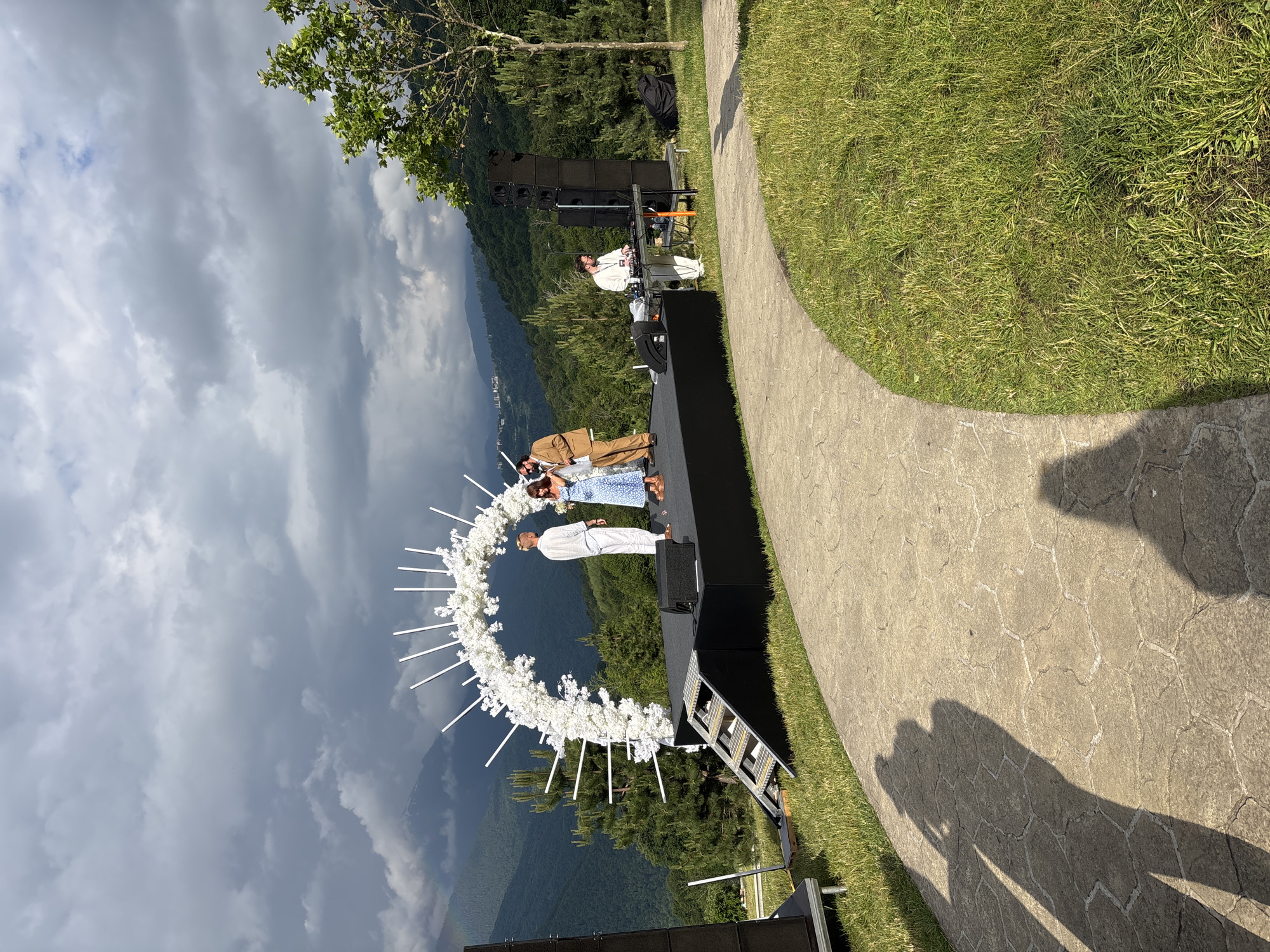 In the city of Estosadok, Russia, an outdoor event is taking place on a grassy hillside with a mountainous backdrop. A stage is set up, adorned with a large, circular archway decorated with white flowers and radiating white spikes. On the stage, three individuals are present: a man in a white outfit, a woman in a blue patterned dress, and a man in a brown suit. The man in the brown suit appears to be interacting with the woman in the blue dress. To the right of the stage, a DJ is set up with a sound system. The surrounding environment consists of lush green grass, a paved pathway, and dense green trees on the slopes of the mountains. The sky is partly cloudy with dramatic storm clouds in the distance, hinting at potential inclement weather. Large speaker towers are positioned on the left and right sides of the stage. Shadows of people are visible on the pathway in the foreground.
