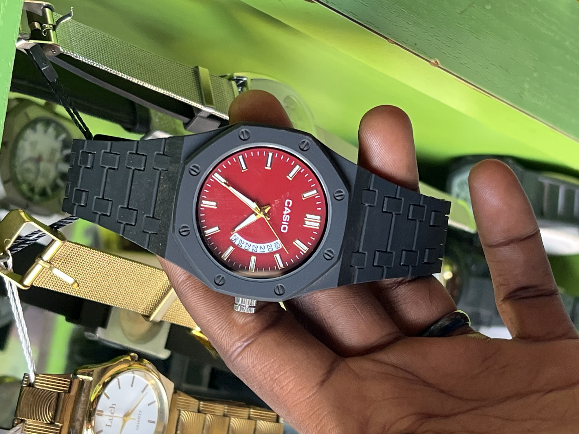 In Bukuru, Nigeria, a close-up, eye-level shot captures a dark-skinned hand holding a black-faced watch with a red dial. The watch's band is made of interlocking black links. In the background, other watches are visible, including one with a gold band and white face, and another with a metallic mesh band. The overall scene appears to be a retail display of watches.