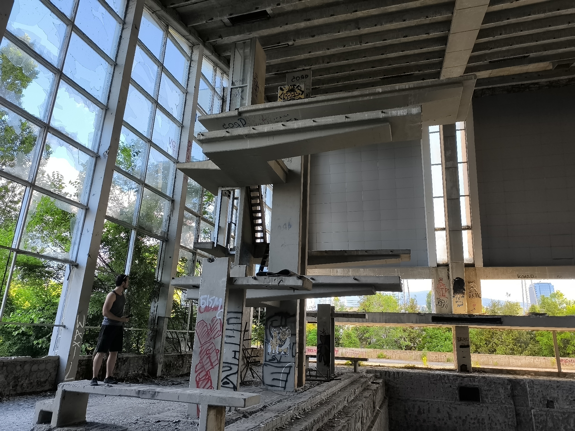 The image depicts the interior of a derelict building, likely an abandoned swimming pool or sports facility, in Sofia, Bulgaria. The structure is characterized by a large, open space with a concrete frame and extensive floor-to-ceiling windows, many of which are broken. Sunlight streams through the windows, illuminating the interior and highlighting the decay.
Dominating the center of the frame is a multi-level concrete structure, possibly a diving platform or spectator seating, covered in graffiti. The graffiti is in various colors and styles, some appearing freshly applied, others faded. A narrow metal staircase is visible leading upwards within this structure.
In the foreground, a man wearing a dark gray tank top and black shorts stands on a concrete platform. He is holding a smartphone in his right hand and looking towards the right side of the frame. His stance suggests he is observing something or taking a picture.
The environment outside the windows is lush with green trees and vegetation, indicating an outdoor setting surrounding the building. In the distance, a modern city skyline is visible, with tall buildings suggesting the proximity of an urban area. The overall impression is one of abandonment and the encroachment of nature into an man-made structure.