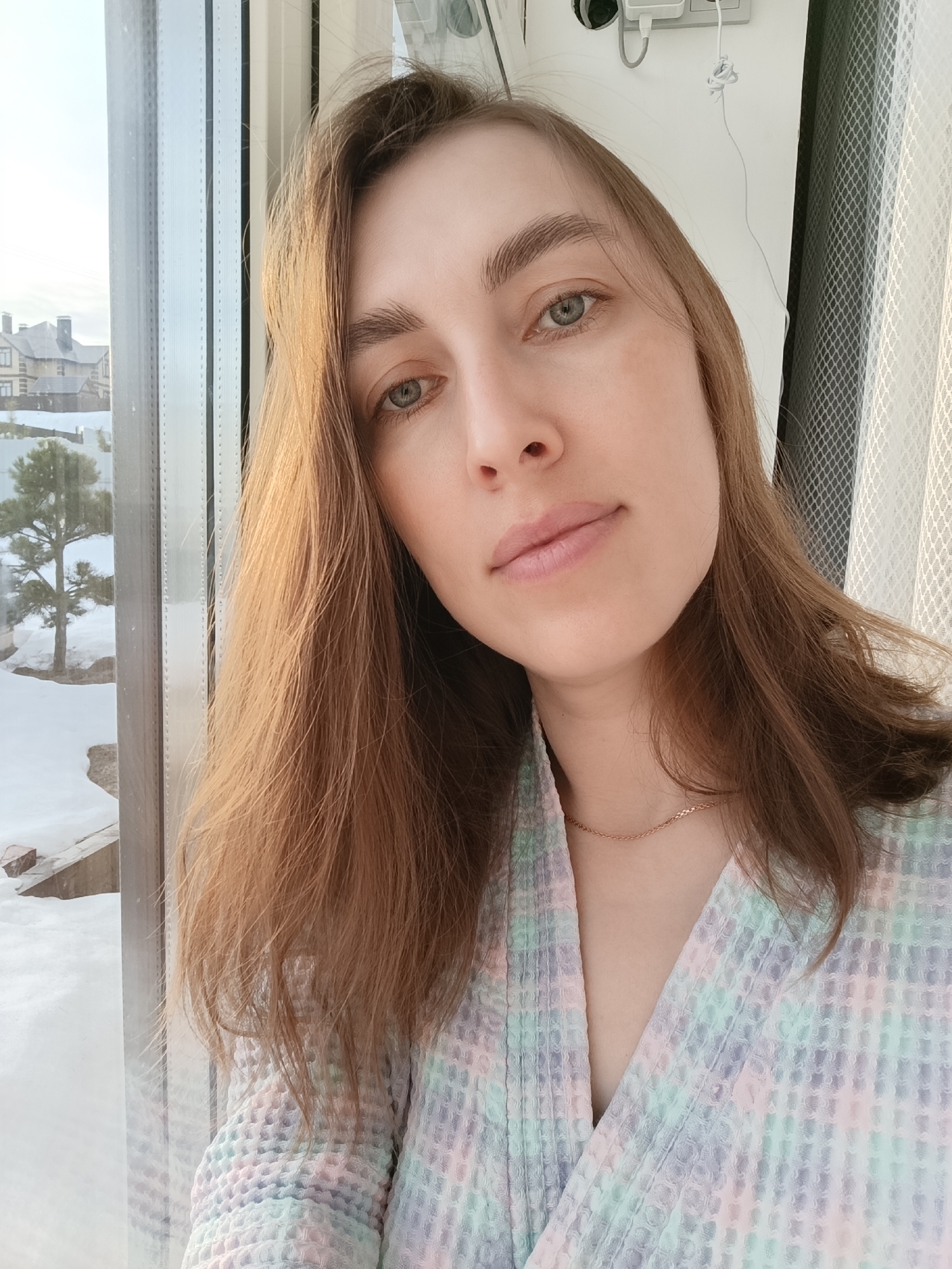 A young adult female is depicted in a medium close-up, facing the camera directly, positioned indoors near a window overlooking a snow-covered residential area in Chaikovsky, Russia. Her long, light brown hair falls over her shoulders. She has fair skin, light-colored eyes (appearing greenish-blue), and a neutral facial expression. A thin gold chain necklace is visible around her neck. She wears a pastel-colored (light pink, green, purple, blue) waffle-knit bathrobe.
To her left, a large window spans vertically, revealing an outdoor scene. The ground outside is covered in white snow. In the middle distance, a dark green coniferous tree stands amidst the snow. Further in the background, a multi-story residential building with a complex dark roof and chimneys is visible. The sky appears bright and clear.
To the right of the woman, a white wall is visible, on which two white electrical outlets are mounted. A white cable is plugged into one outlet, and another white cable, partially knotted, hangs below. A sheer, white, mesh-patterned curtain is partially visible on the far right edge of the frame.