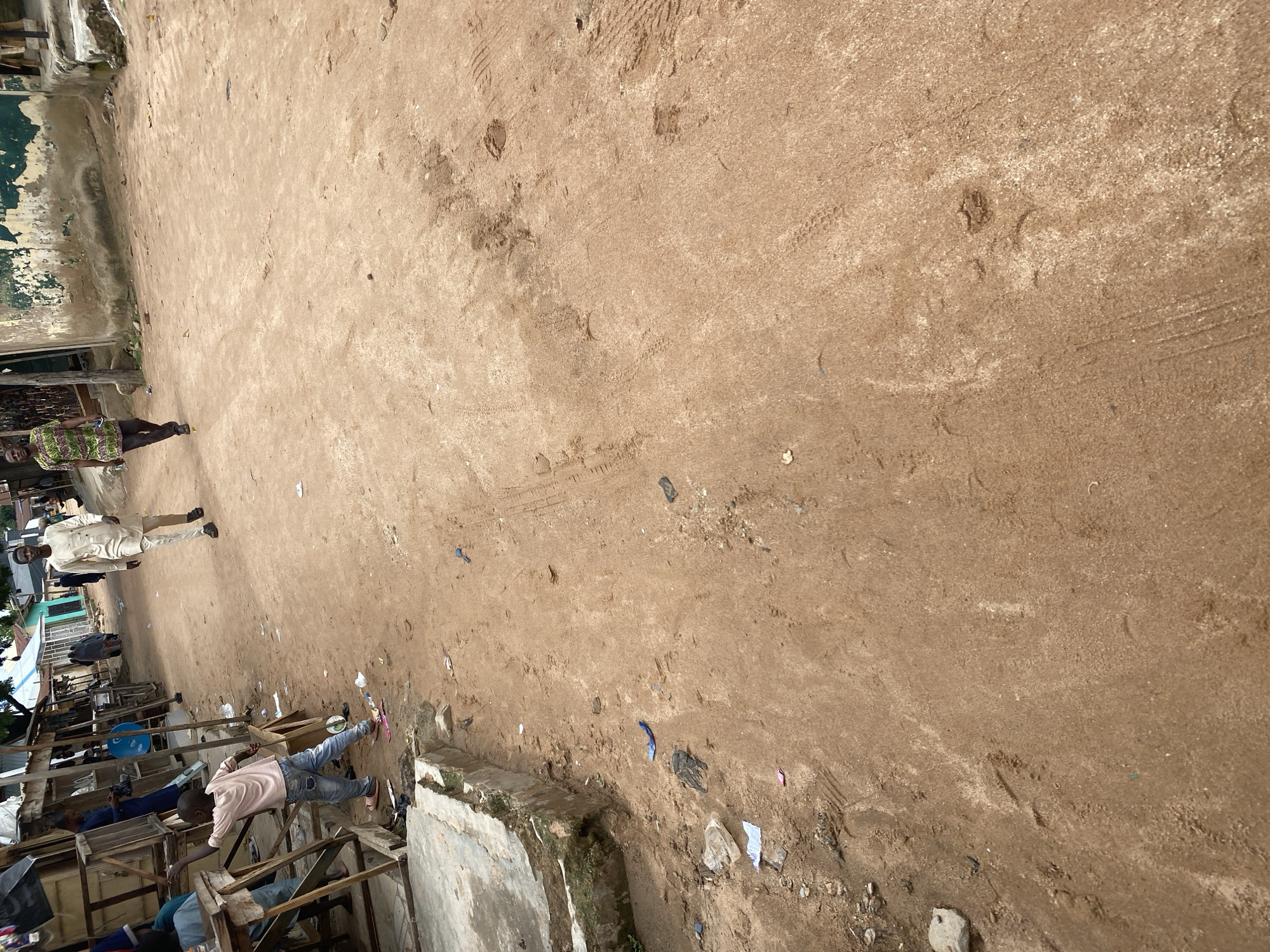 The image depicts a dusty, unpaved street in Jalingo, Nigeria, likely a marketplace or residential area, characterized by makeshift stalls and buildings. In the foreground, a young child in a pink shirt and blue jeans is interacting with a wooden stall. Further down the street, two men are walking away from the camera. One man is wearing a light-colored traditional outfit, and the other is in a colorful patterned shirt and dark trousers. The ground is composed of loose, reddish-brown soil with tire tracks and scattered debris, including plastic waste. The environment appears warm and dry, typical of the region.