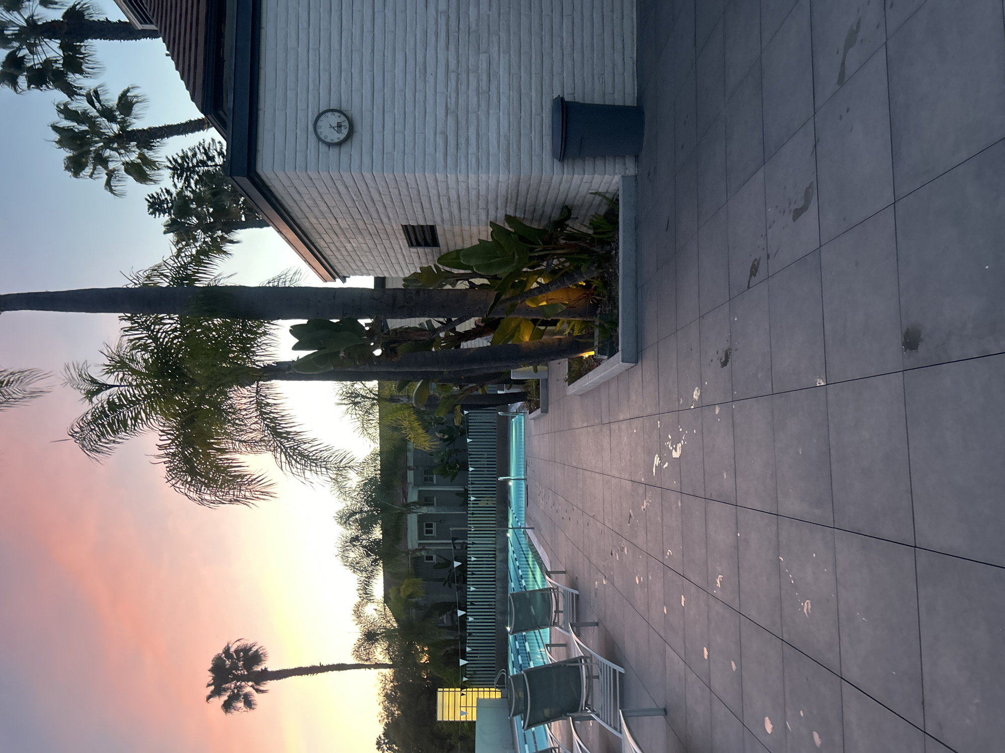 The image depicts a poolside scene at dusk in Los Angeles, United States. The environment features a swimming pool with lanes marked by buoys and lounge chairs arranged alongside the pool deck. Palm trees are silhouetted against a sky displaying colors of pink and orange, indicating either sunrise or sunset. A building with a white brick exterior is present in the background. The pool deck is tiled and shows some signs of water splashes. A clock is visible on the building's exterior. A trash receptacle is located near the building.