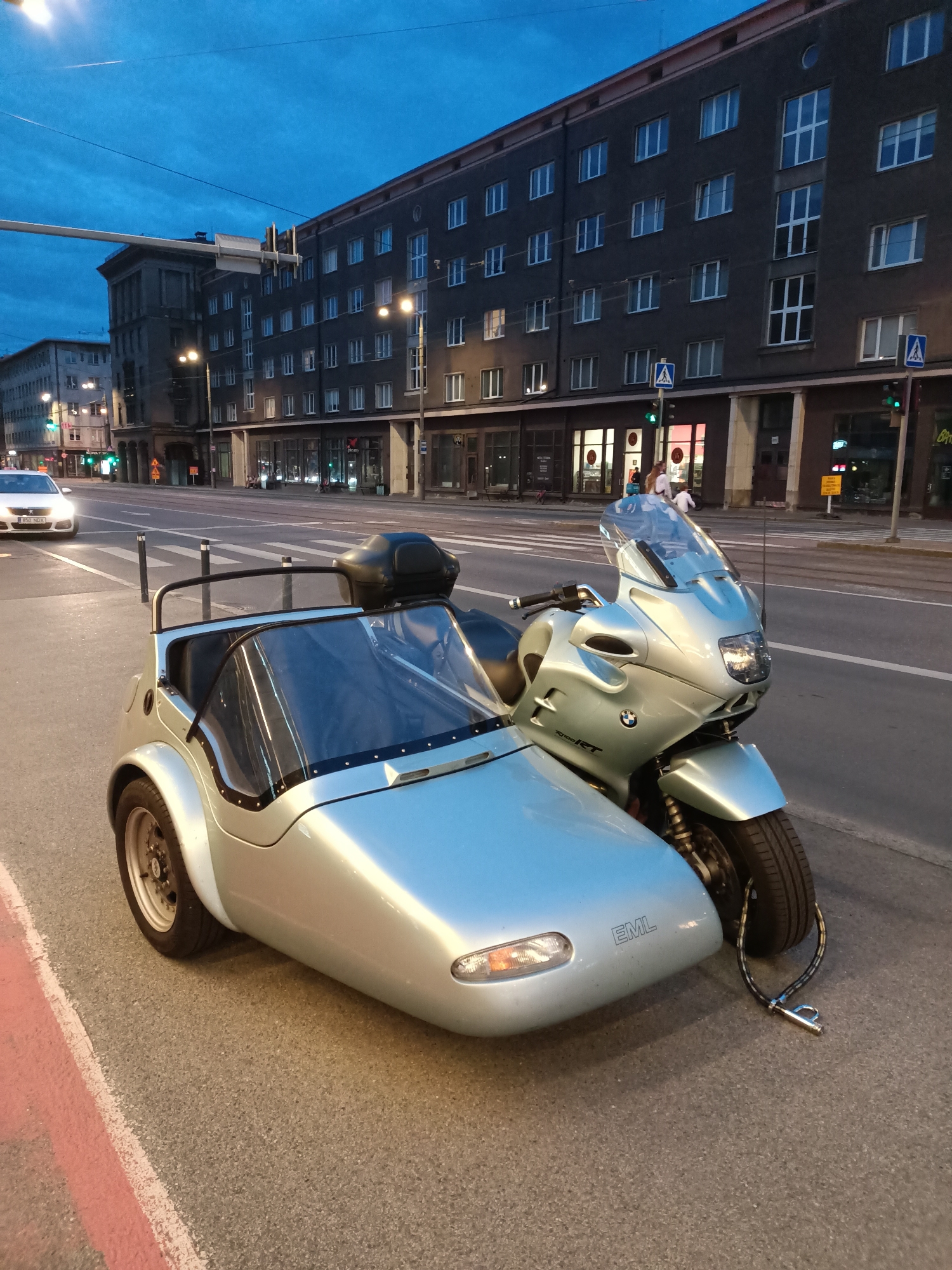 A silver BMW motorcycle with a matching sidecar is parked on the right side of a city street in Tallinn, Estonia. The motorcycle has a tall windscreen and a black storage box on the rear. The sidecar is a light blue color and has a small windshield. A black cable lock is attached to the front wheel of the sidecar and the motorcycle's frame. The street appears to be a main thoroughfare, with multiple lanes of traffic and a pedestrian crossing in the foreground. Buildings with many windows line the street, suggesting an urban environment. The sky is a deep blue, indicating dusk or twilight. No people are directly interacting with the motorcycle or sidecar. A white car is visible in the distance on the left side of the road.