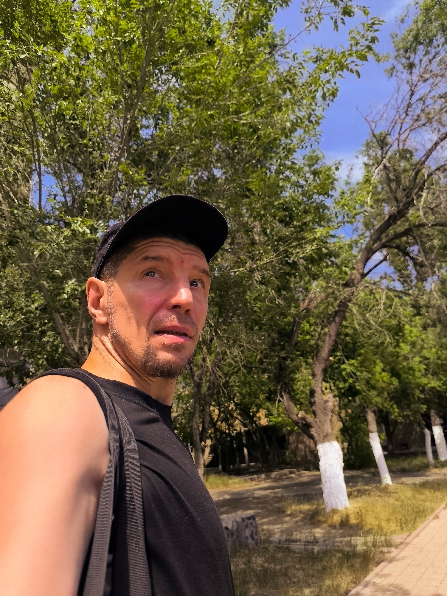 A medium shot, selfie-style photograph captures a man wearing a black baseball cap and a black tank top, outdoors on a sunny day. The man has fair skin, short brown hair, a light brown goatee, and his eyes are looking upwards and to the right. He is positioned in the foreground, with lush green trees and the blue sky forming the background. Some of the tree trunks in the background are painted white. The man's right arm and shoulder are visible on the left side of the frame. The scene appears to be in a park or a tree-lined area within the city of Karaganda, Kazakhstan.