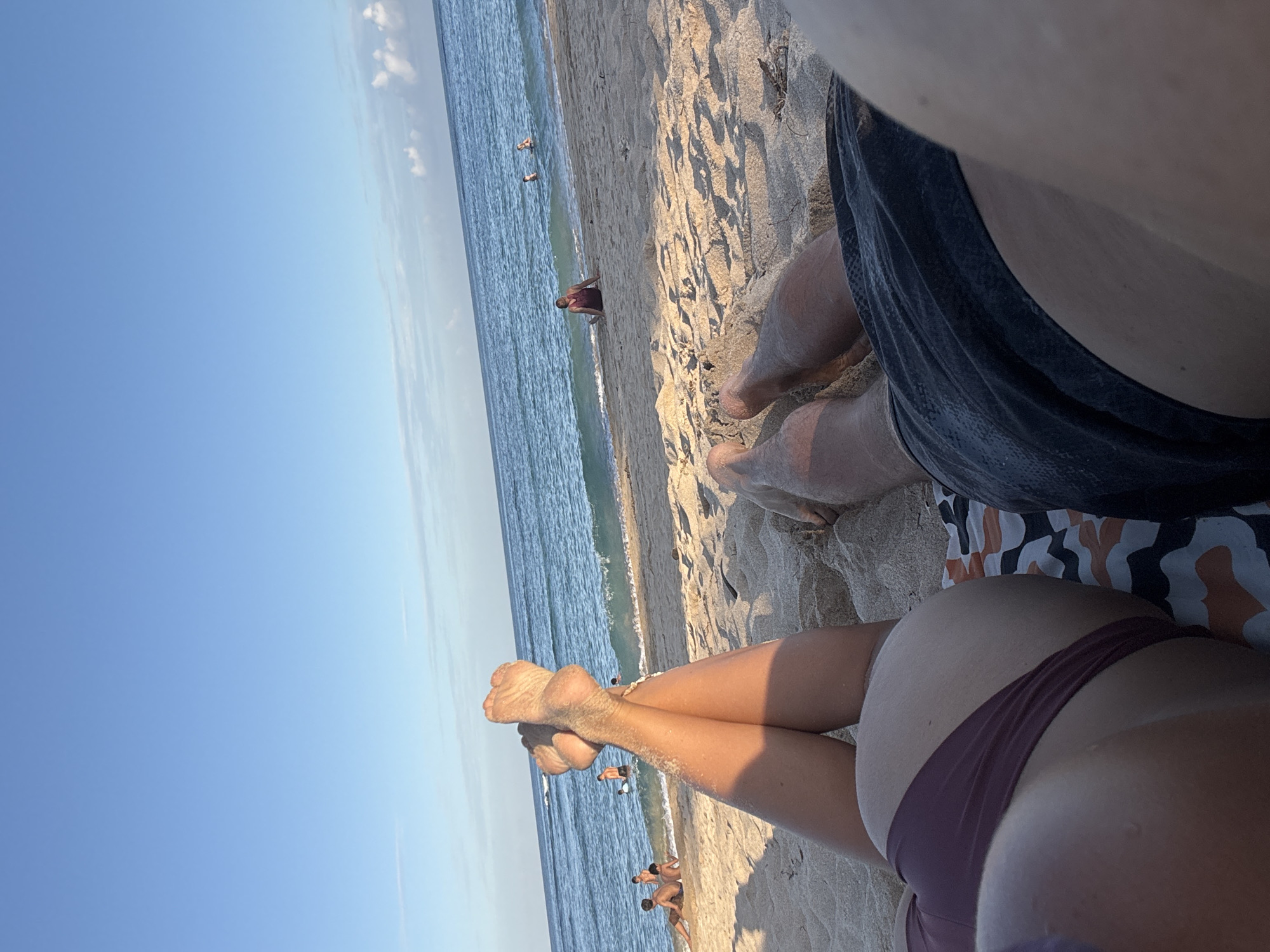 The image shows a beach scene with a view from a person lying on the sand in Kuta Selatan, Indonesia. The sky is clear blue. In the distance, people are visible in the water. In the foreground, the legs of two individuals are visible; the closest person is wearing a bathing suit, while the other is wearing shorts.