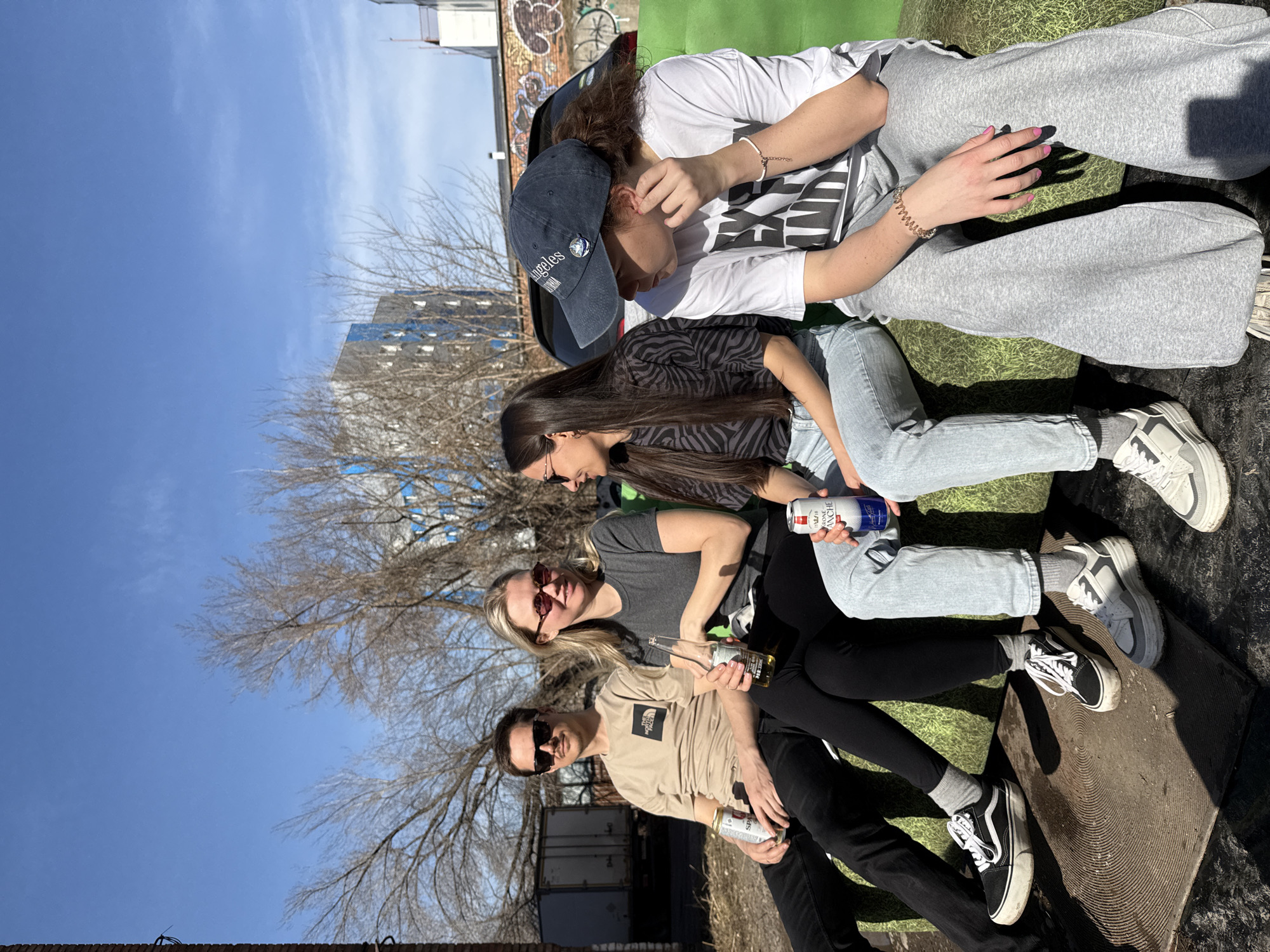 Four individuals are seated outdoors on a surface made of metal near what appears to be containers; one can see graffiti in the background. The person seated closest to the camera is a man with dark hair wearing sunglasses, a beige shirt, and black pants, and holding a beverage can. Adjacent to him sits a blonde woman with sunglasses, wearing a grey shirt. Next to her is a woman with long brown hair and sunglasses, wearing a black-and-white patterned shirt. The individual farthest from the camera is wearing a white shirt, grey sweatpants, and a dark-colored baseball cap. The individual has their hand near their face.
The sky is visible and blue, with some trees that do not have leaves yet. The picture was likely taken during the spring or fall season. The backdrop includes an urban area with buildings.
It is plausible that the picture was taken near the location called Krasnoyarsk.