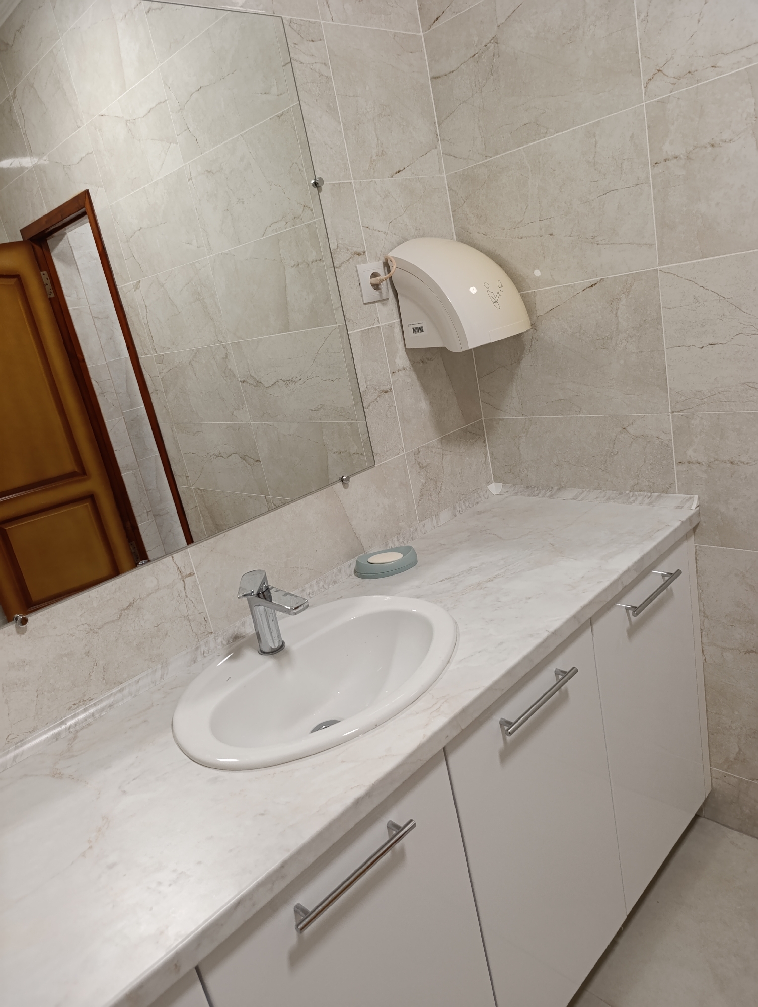 The image shows a modern bathroom sink area, likely located within the Dubăsari town council building in Moldova. A white ceramic sink with a chrome faucet is mounted on a white marble-effect countertop. Below the countertop are white cabinets with chrome handles. To the right of the sink, a white automatic hand dryer is mounted on a tiled wall. A mirror above the sink reflects a wooden door on the left side of the frame. The walls are covered with light-colored marble-patterned tiles. The floor appears to be tiled with a neutral color.