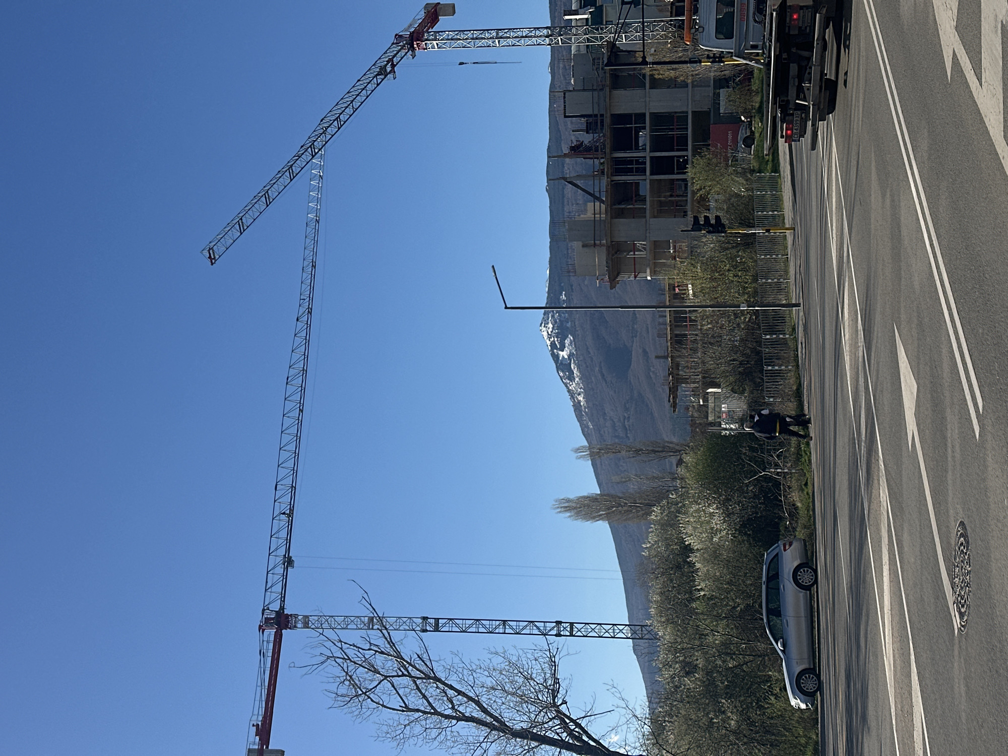 The image shows an urban scene with several prominent features, including two large construction cranes, a multi-story building under construction, a roadway with white lane markings, a mountain range with snow-capped peaks in the background, and a clear blue sky.
A silver car is parked along the roadside. A person wearing a dark jacket and light-colored pants is walking alongside the road. A light pole and traffic lights are also visible.
The image is taken in Sofia, Bulgaria.