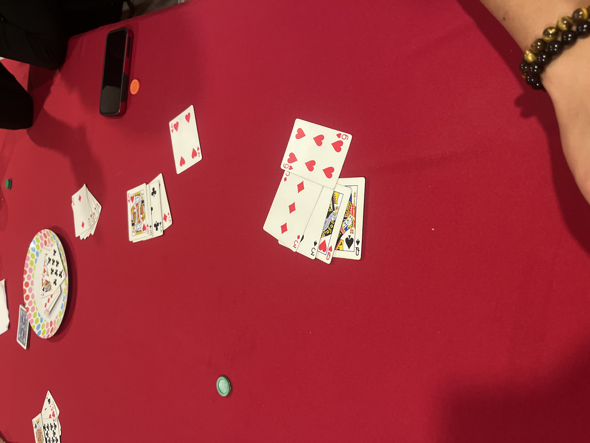 The scene is indoors on a red surface, possibly a table. There are playing cards scattered around. Some cards are stacked in groups, while others are isolated. There is a phone, a paper plate with more cards on it, and poker chips present. A person is visible in the upper right wearing a beaded bracelet. Barrio de Nuevo León, Mexico is not discernible from the image.