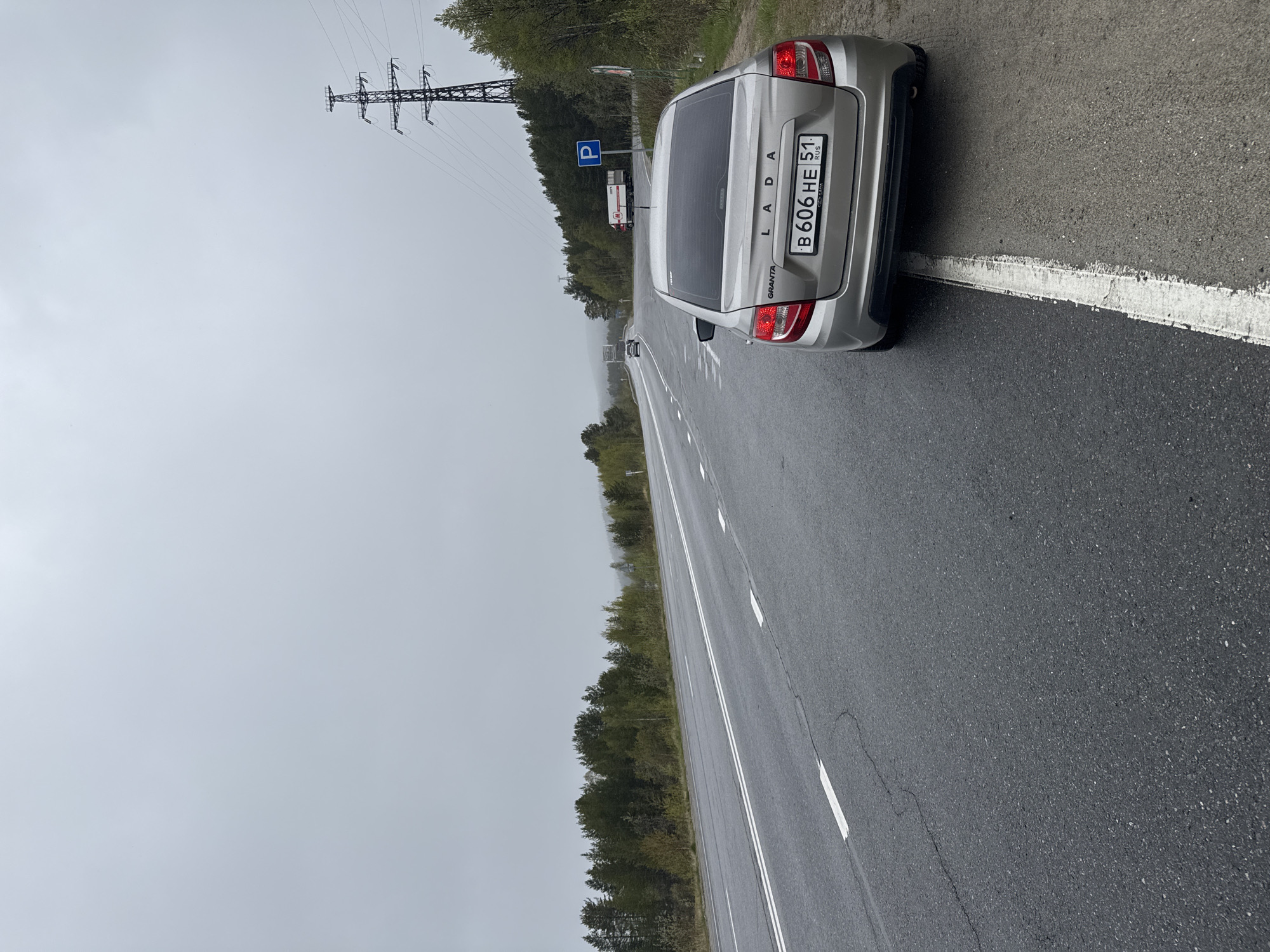 The image depicts a silver Lada Granta parked on the shoulder of a wet asphalt road. The license plate reads "B 606 HE 51 RUS". The road has white lane markings. A power transmission tower is visible in the distance. Trees line the roadside. A "P" parking sign is also visible. The sky is overcast. The general location appears to be a roadside area near a forested region. There are a few vehicles ahead on the road, one possibly a truck. Based on the license plate information, the location is likely near Tik-Guba, Russia.