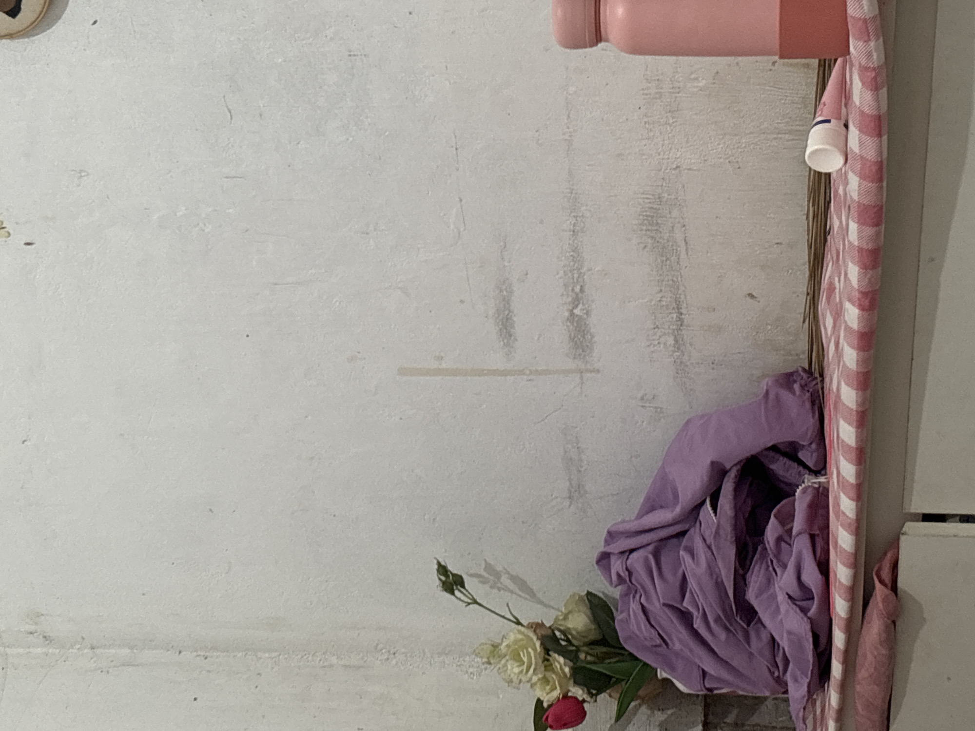 The image shows a close-up of a shelf in what appears to be a domestic setting, likely a home. The shelf is covered with a pink and white checkered fabric. On the shelf, to the left, is a bouquet of artificial flowers, including white roses and a single red tulip, placed in a vase. A crumpled piece of purple fabric is draped over the flowers and the shelf. To the right of the flowers, a small broom made of twigs rests on the shelf. Further to the right, a pink water bottle is visible, and next to it, a small white tube with a blue band and a white cap lies on its side. The background is a rough, off-white textured wall with some faint grey marks and a vertical faint line. The overall impression is of a simple, cluttered shelf, possibly in a room in North Jakarta, Indonesia, though this cannot be definitively confirmed from the visual information alone.