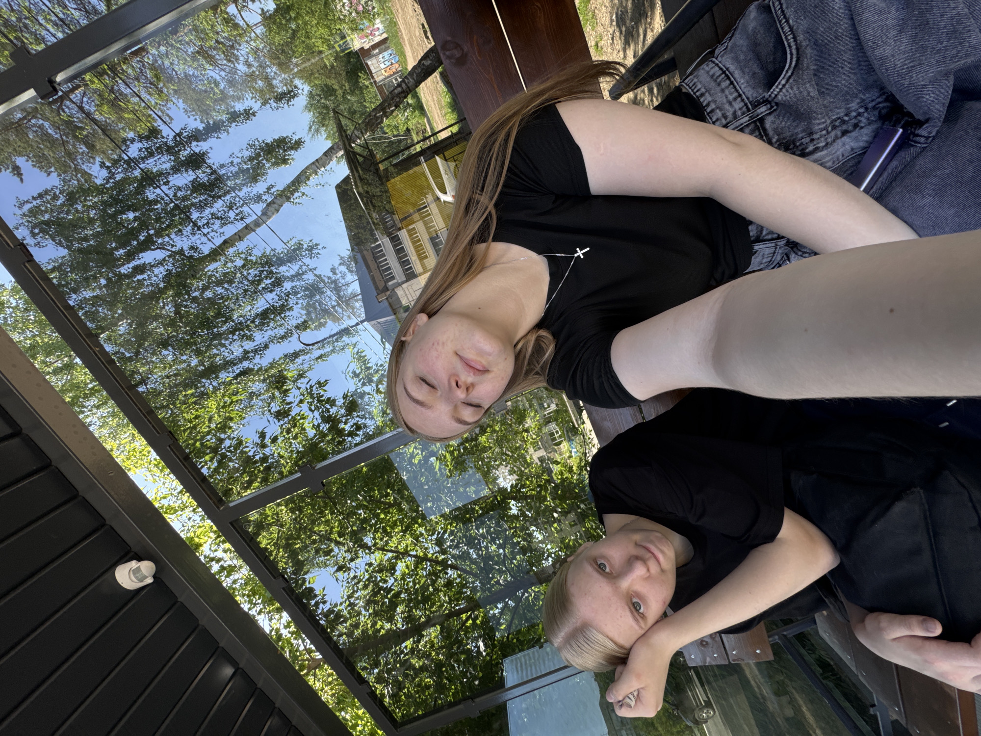 Two young women are sitting on a bench under a glass-roofed structure. One has long brown hair and is wearing a black t-shirt with a cross necklace. The other has short blonde hair and is wearing a black t-shirt. They appear to be in Chaikovsky, Russia, based on the reflection in the glass roof, which shows buildings and trees typical of the area. A wooden bench and a pair of grey jeans with a purple vape are visible. The glass roof reflects the sky and trees.