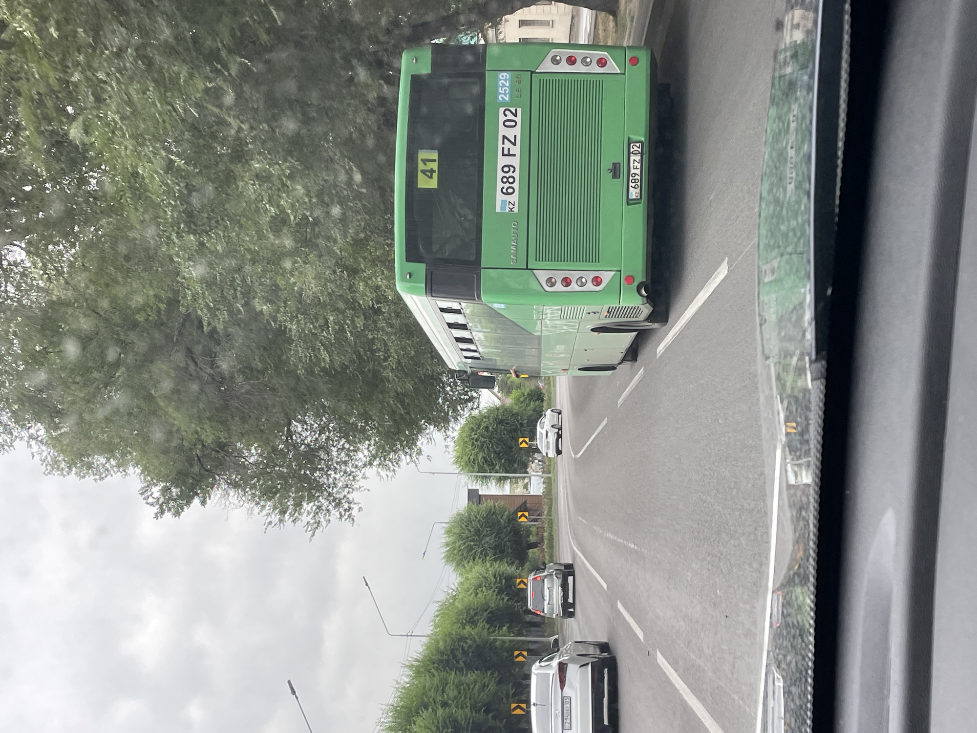 The image shows a street scene in Almaty, Kazakhstan, likely on a cloudy day, with a green bus in the foreground. The bus has the number "41" displayed on its rear window and license plate "689 FZ 02". Several cars are visible on the road, including a white sedan with license plate "214 XF 05" and a silver SUV. The street is lined with manicured trees and streetlights. The perspective suggests the photo was taken from inside another vehicle.