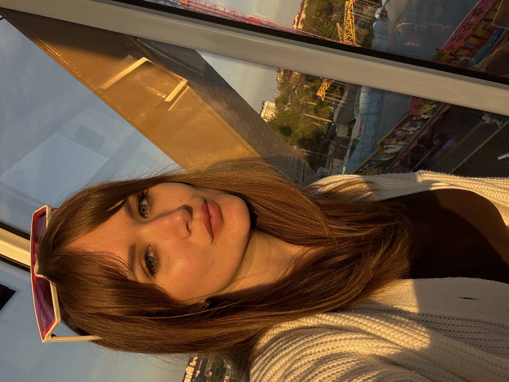 A sideways medium shot depicts a fair-skinned woman with long brown hair and green eyes. Her hair has bangs and hangs loose around her face, one side adorned with pink-framed sunglasses. She wears a white knit sweater over a brown top. The background, visible through a window frame, features a cityscape with buildings, trees, and amusement park rides. It is sunset. The scene takes place within a moving ferris wheel cabin. This photograph appears to have been taken in Yevpatoriya, Ukraine.