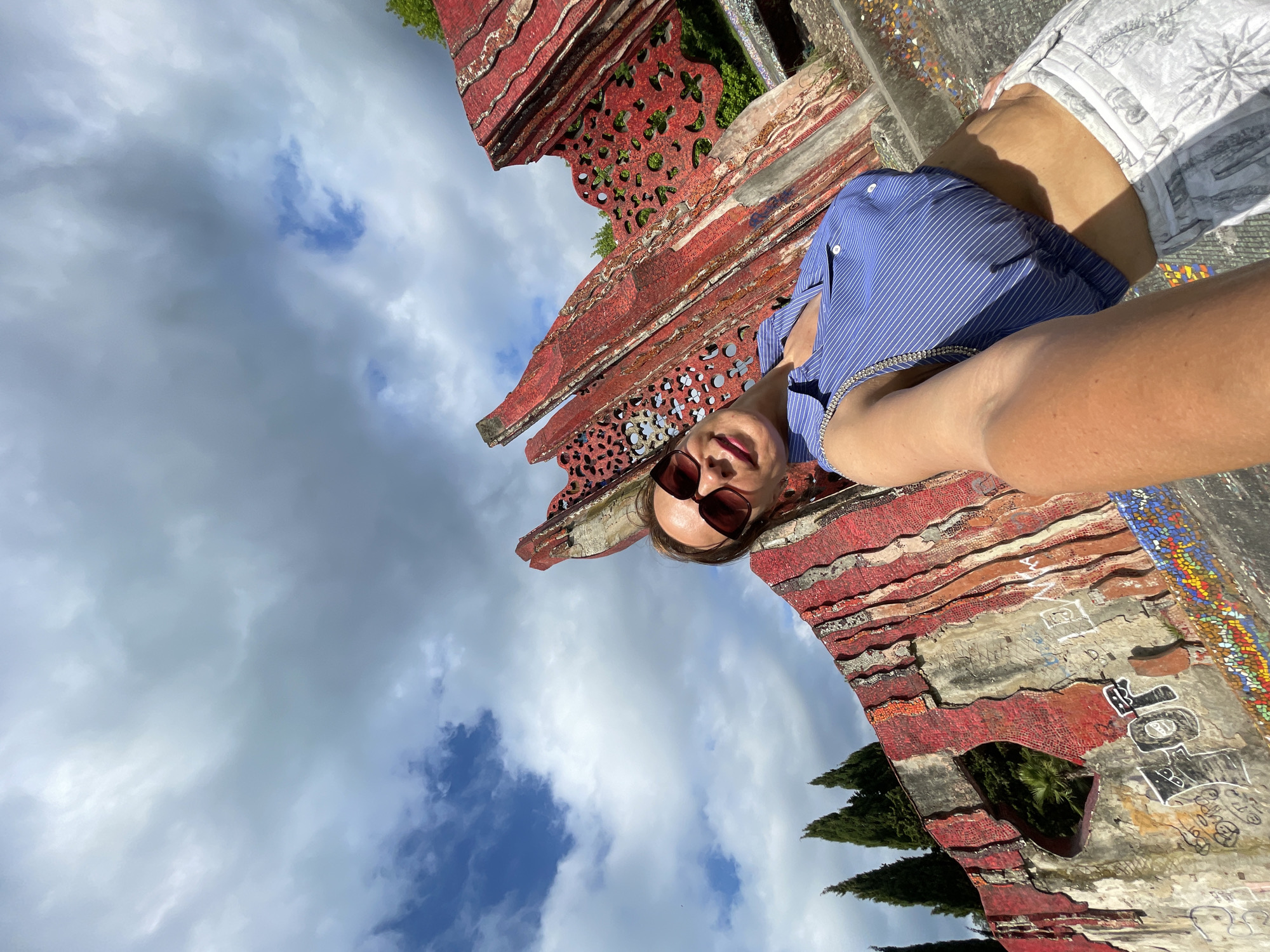 A woman is lying down, taking a selfie in front of a red, ornate structure with intricate patterns and mosaic details. She is wearing a blue and white striped shirt, white shorts with a compass design, and sunglasses. The sky is cloudy with patches of blue. The structure appears to be part of a building or a garden feature, possibly an archway or a decorative wall. Some graffiti is visible on the structure. The scene suggests a sunny day, possibly during the daytime, given the lighting. The woman's expression is relaxed, and the overall mood is casual and cheerful. The location is identified as Sochi, Russia.