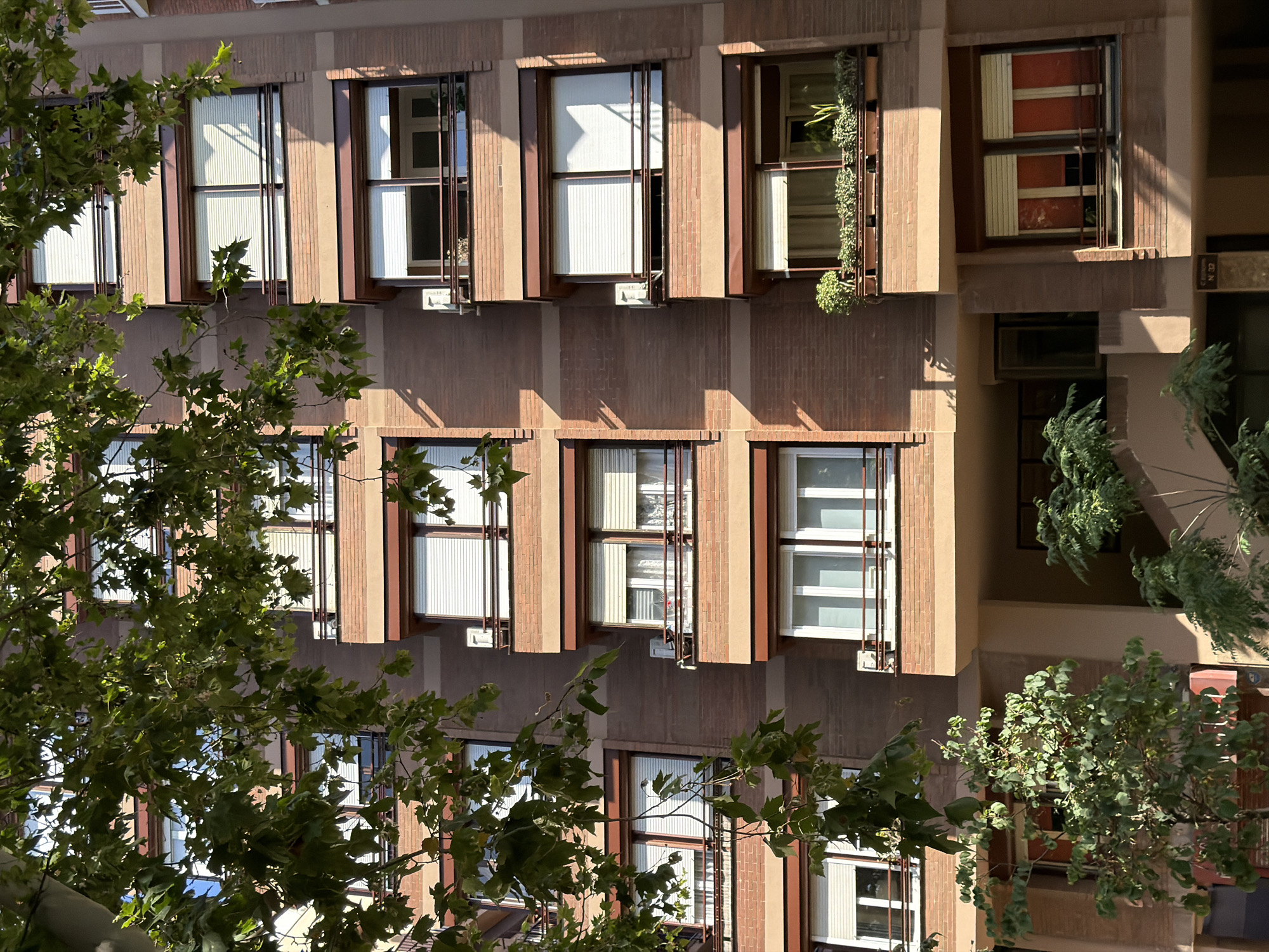 The image shows a building exterior with multiple windows, some open, some closed, and some adorned with plants. The building appears to be multi-story, with a facade of brown and beige bricks and vertical decorative elements. Sunlight casts shadows on the building, suggesting it is daytime. The windows have a distinctive design with vertical metal bars and blinds. Some windows have plants growing in planters outside them. The presence of lush green foliage in the foreground, extending into the frame, indicates a natural element integrated with the architecture. The overall impression is of an urban setting, possibly with a focus on green architecture or well-maintained outdoor spaces. The specific location cues of Barcelona, Spain, combined with the architectural style, might suggest a Mediterranean influence. There is no visible text in the image. The scene is calm and serene, with the interplay of light and shadow creating a visually interesting composition. The focus is on the building's architecture and its interaction with nature.