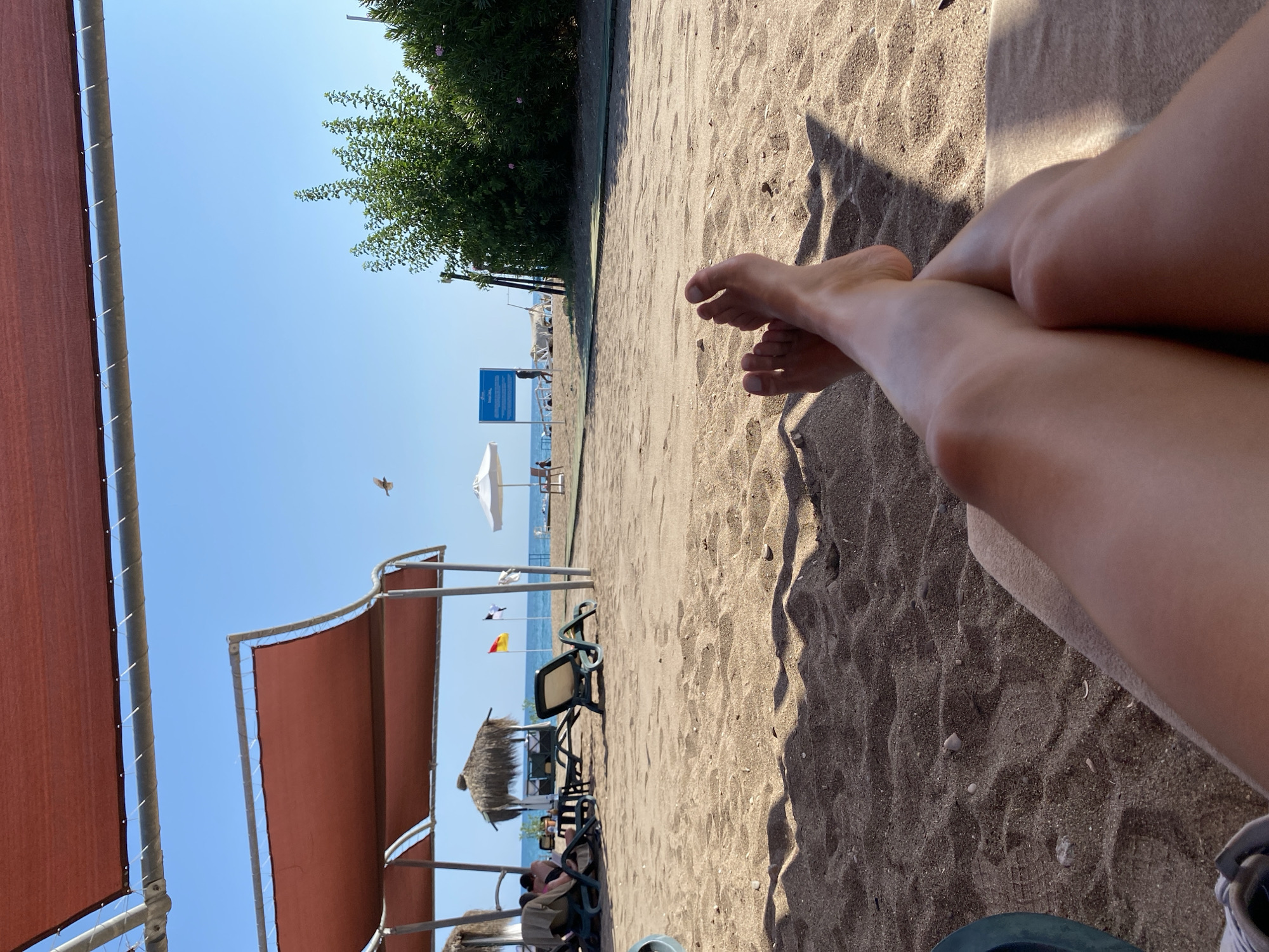 The image shows a first-person perspective of someone relaxing on a sandy beach in Aksu, Turkey. The foreground is dominated by the person's legs and feet, crossed at the ankles, resting on a towel or lounge chair, with the sand in sharp focus. In the mid-ground, the beach stretches out, with several empty lounge chairs, some with towels, under a reddish-brown awning structure. Further back, the turquoise sea is visible, with a white umbrella and a few figures walking near the water's edge. A sign with blue and white text is also present. The background features lush green bushes and trees, and a clear blue sky. The overall scene depicts a serene beach environment.