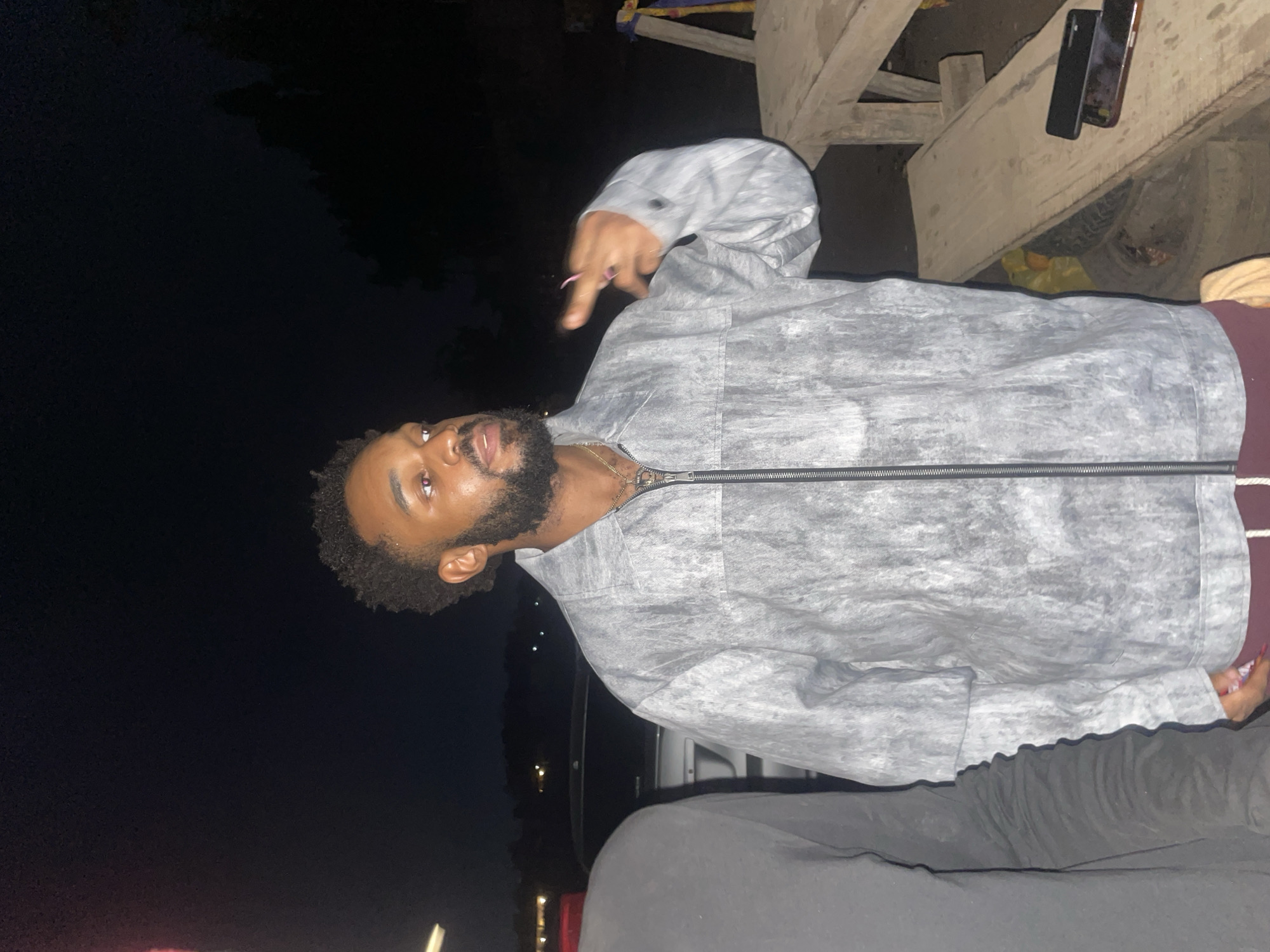 A person with dark skin, a beard, and dark curly hair is wearing a gray jacket and black pants. The person is giving a hand gesture. Two phones are visible on a wooden surface above the person. The location is outdoors at night, possibly in Galadimawa, Nigeria.
