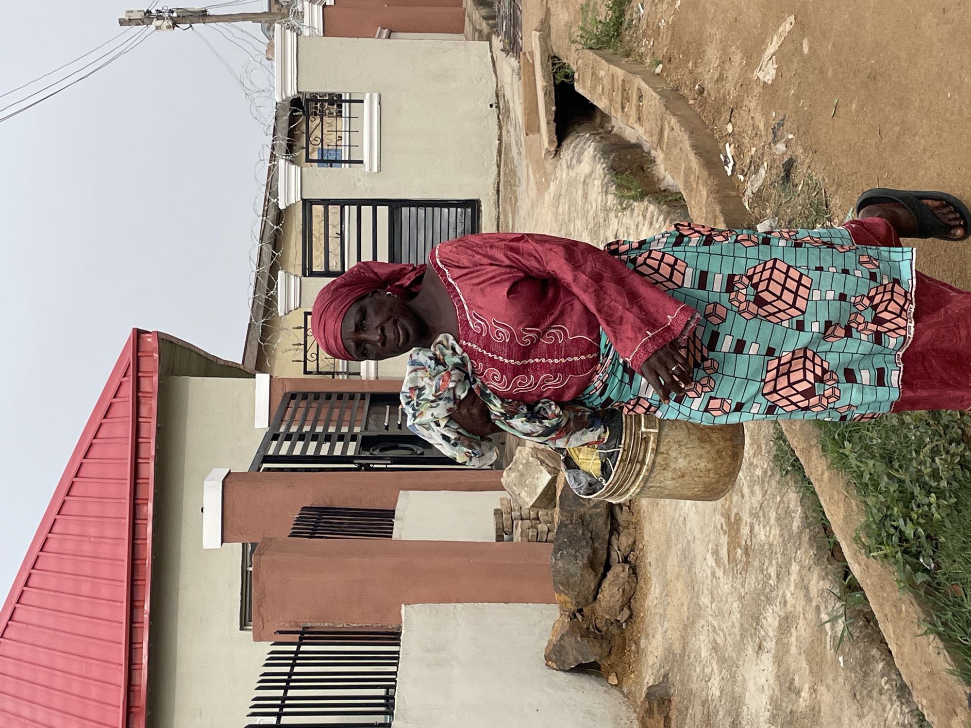 An elderly African woman is standing outside on a concrete walkway in Jos, Nigeria. She is wearing a red headscarf, a red long-sleeved blouse with white trim, and a turquoise wrap skirt with a geometric cube pattern. She is holding a large bucket. To her left is a building with a red roof and tan walls. The building has black bars over the windows. Above the building is a power line. To her right is a concrete drainage ditch. The ground is dirt and concrete. The sky is overcast.