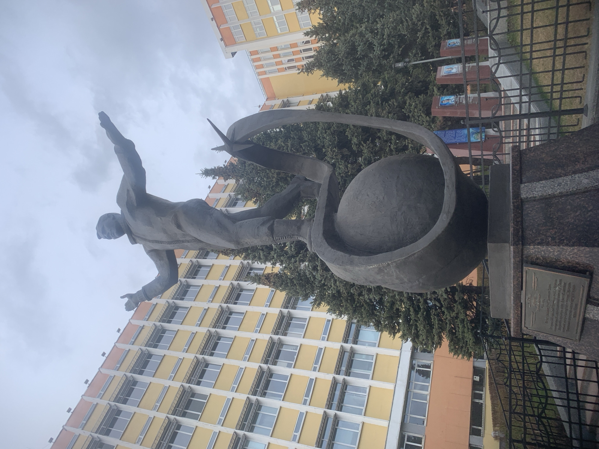 The image shows a statue in Izhevsk, Russia. The statue depicts a man seemingly leaping above a globe encircled by an abstract, ribbon-like structure. A small, metallic plaque is affixed to the statue's base. In the background, a tall building with a yellow facade and rows of windows is visible. A fence separates the statue from a row of memorials. Trees provide a partial backdrop to the statue. The sky appears overcast.