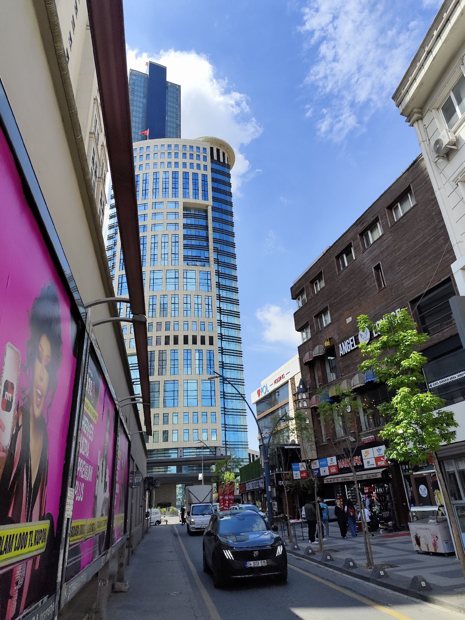 Here is a detailed description of the image:
**Setting:** An urban street scene in the city of Unknown, Unknown. The viewpoint is at street level, looking down a narrow road with buildings on either side.
**Architecture:** The left side of the road is lined with buildings featuring advertising billboards. Across the street on the right are shops and a building with a wooden facade advertising “ANGEL LOUNGE”. Further back, there is a modern high-rise with blue windows.
**Transportation:** A dark-colored car is driving down the road, license plate visible: "34 BTU 519". Further in the background, a truck is seen.
**People:** Pedestrians are visible walking along the sidewalk on the right side of the street, looking at shops.
**Objects and Details:** Advertising billboards dominate the left side of the road. Small trees have been planted along the sidewalk on the right. The sky is blue with scattered clouds.