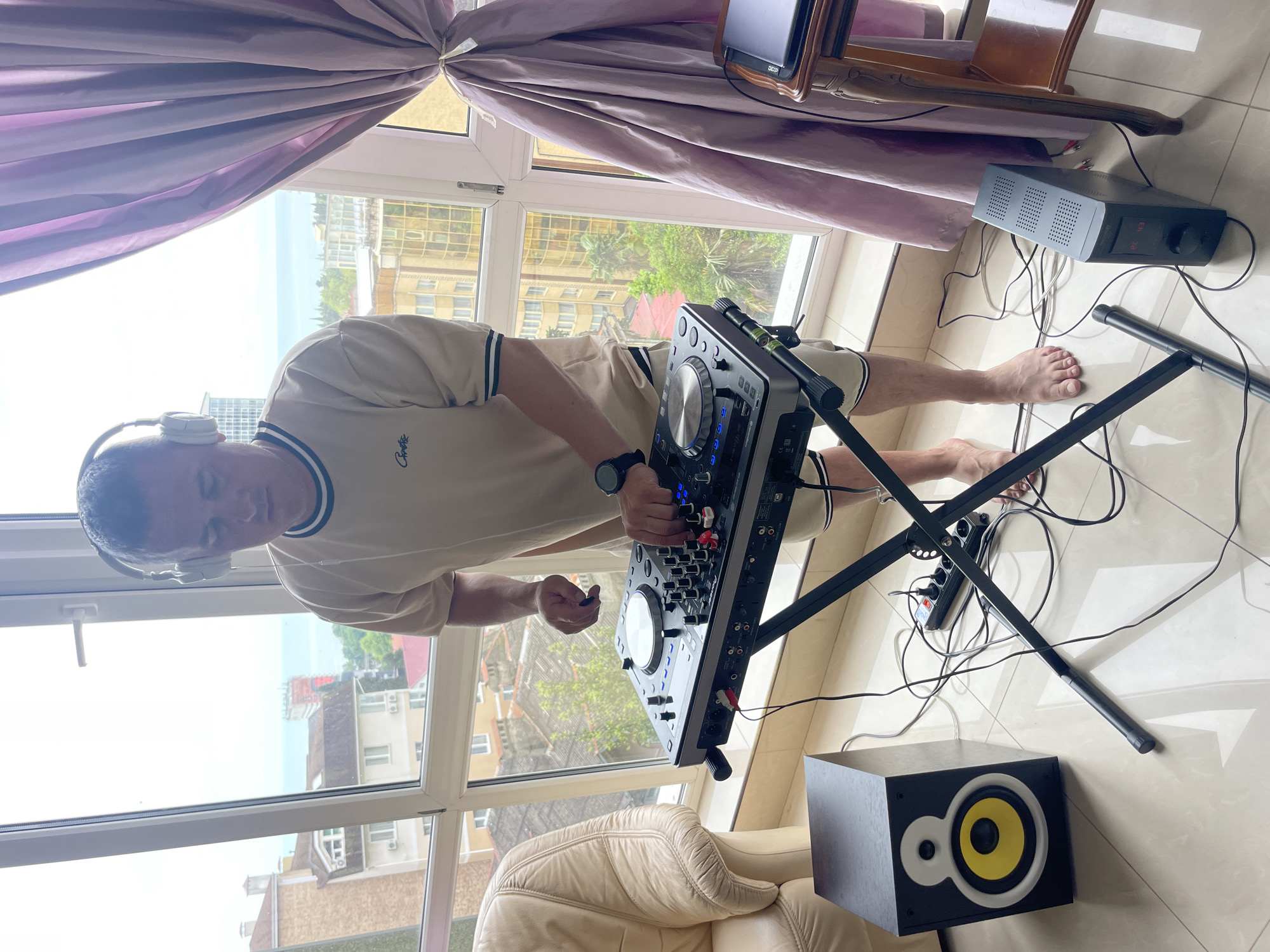 A medium, indoor shot depicts a man standing barefoot in front of a DJ console set up on a stand. The man has headphones on and appears to be operating the console. A black box-shaped object with a yellow circle and white ring is on the floor. There is another electronic device sitting on the floor behind the console stand near a window. The shot is angled to show a cityscape in the background, and based on available clues, the location is potentially in Sochi, Russia. A light purple curtain is partially visible above the window. A beige leather armchair is present in the bottom left corner.