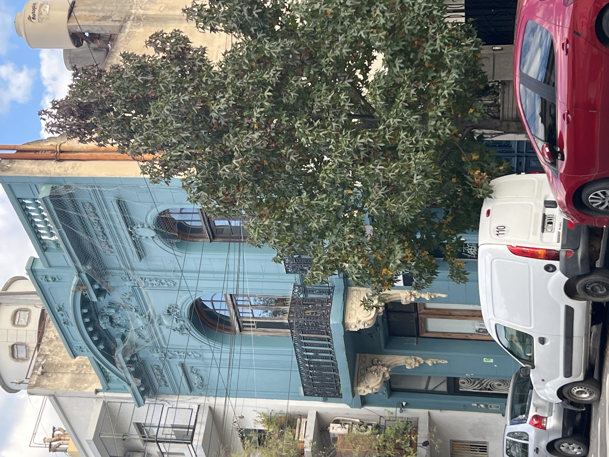 A vertical eye-level outdoor shot in Buenos Aires, Argentina, reveals an ornate, light-blue building partially obscured by a large tree and two parked vehicles. The building, possibly a residence or commercial structure, is characterized by detailed molding, decorative flourishes, and arched windows with ornate balconies. A tree with dense green foliage dominates the center, concealing parts of the building's facade. Two vehicles are parked in front of the building: a red car to the right and a white van slightly behind it. Another vehicle is partially visible near the white van. The buildings and street infrastructure are in moderate condition with possible aging or wear. The sky above is slightly overcast with a pale blue hue.
