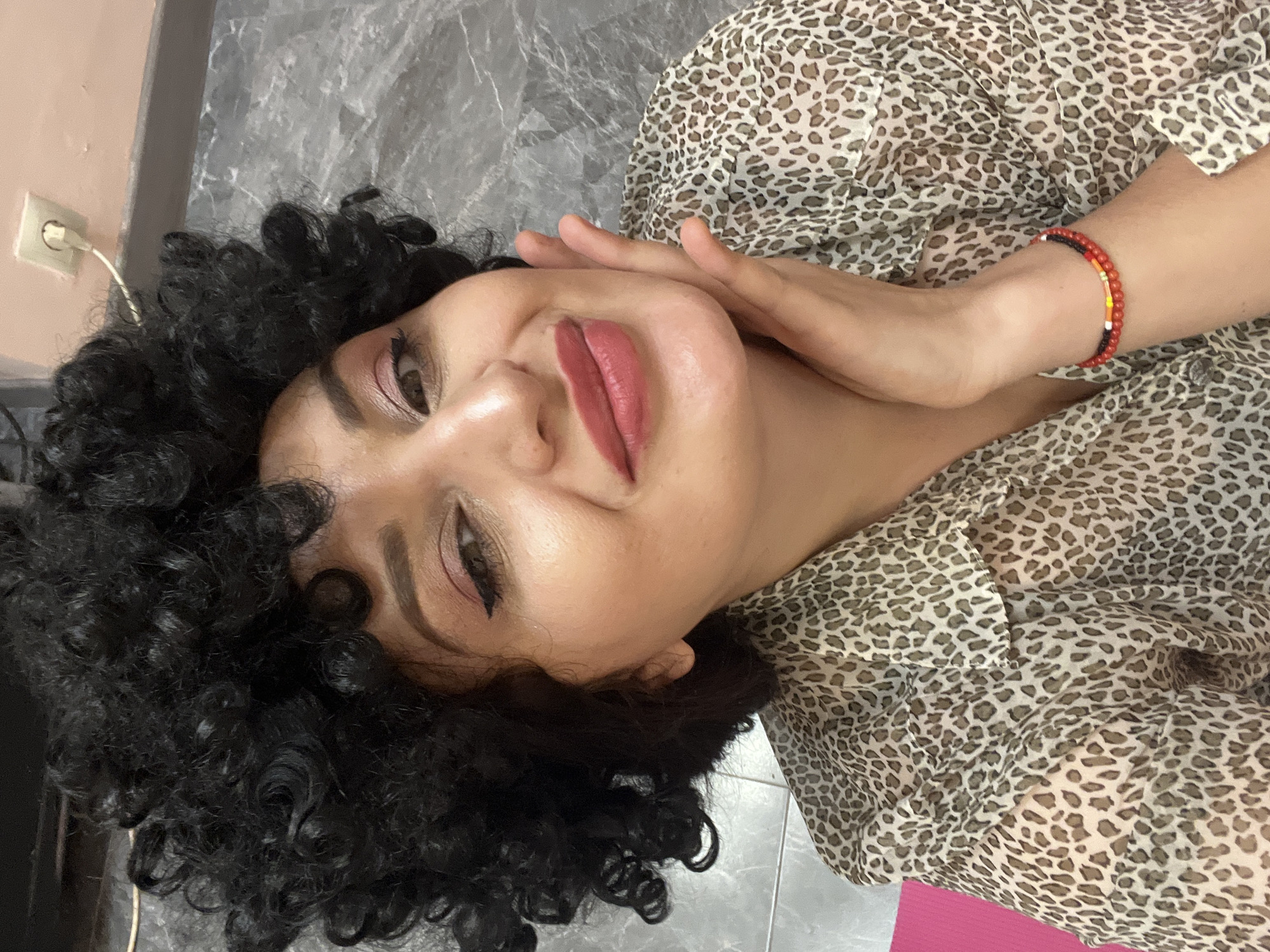 A head-and-shoulder portrait captures an adult female with light to medium skin tone, black curly hair, and brown eyes, located in the city of Unknown, Unknown. She presents a smiling expression with her head slightly tilted to her left, her right hand gently resting against her right cheek and jawline. Her eyes feature black winged eyeliner and pink eyeshadow, complementing her reddish-pink lipstick. She is attired in a leopard-print collared shirt. A multi-colored beaded bracelet, composed of red, black, yellow, and white beads, is visible on her right wrist. The background features a light pink wall with a white electrical outlet and plug in the upper right. Below this, a dark grey strip separates the pink wall from a mottled grey surface. A bright pink mat or object is partially visible on the lower left side of the frame.